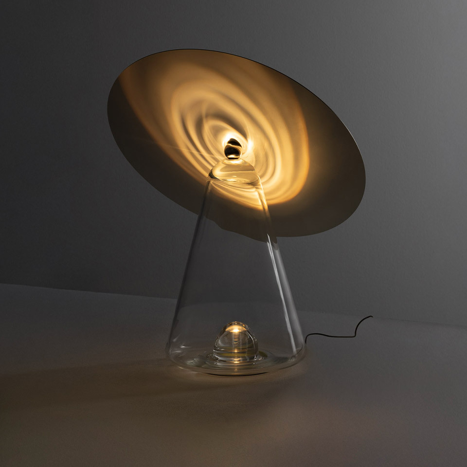 Artemide Boltons table lamp: tilted gold disc reflector on clear conical glass base, casting warm spiral light patterns
