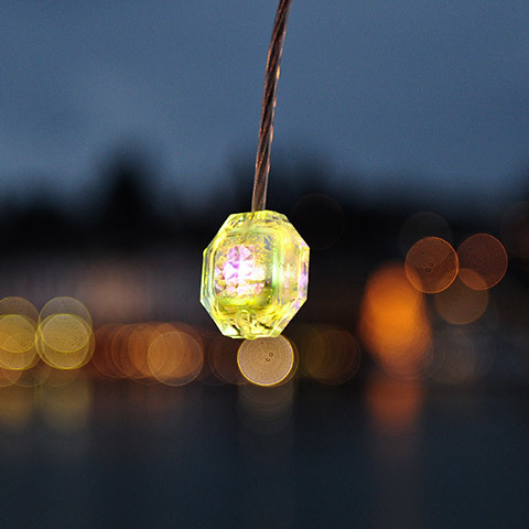 Lampada a sospensione Diamante: piccola lampada geometrica sfaccettata simile a un cristallo, luminosa giallo-verde, sospesa su un cavo con sfondo bokeh al crepuscolo