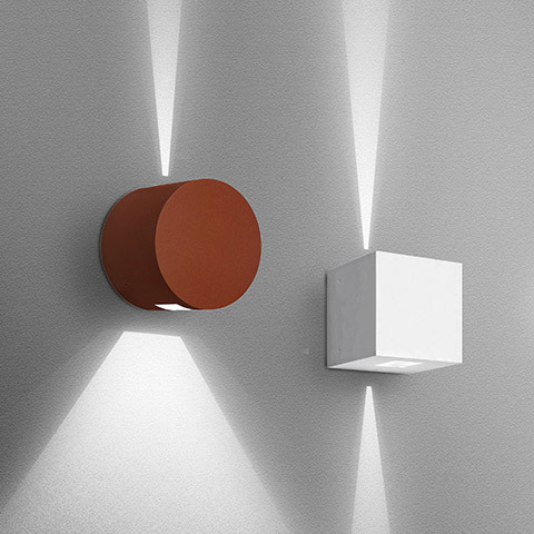 Due applique Artemide Effetto: cilindrica rosso ruggine e cubica bianca, entrambe proiettano fasci di luce verso l'alto e il basso su parete grigia