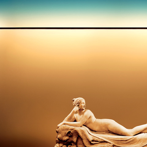 Artemide Dusk linear lamp casting warm golden light over a classical reclining female sculpture on a lion, teal sky above