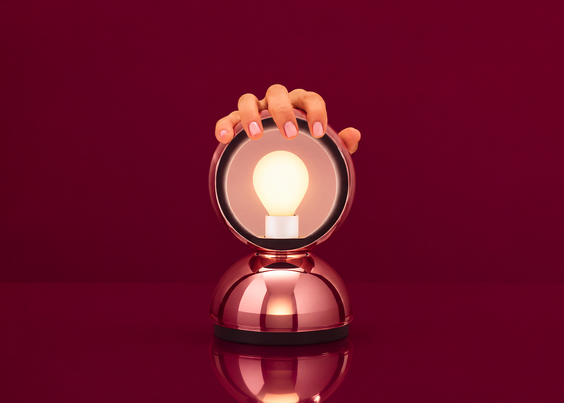 Mano che tocca una lampada da tavolo oro rosa con cornice circolare a specchio e lampadina Edison accesa su superficie riflettente, sfondo rosso