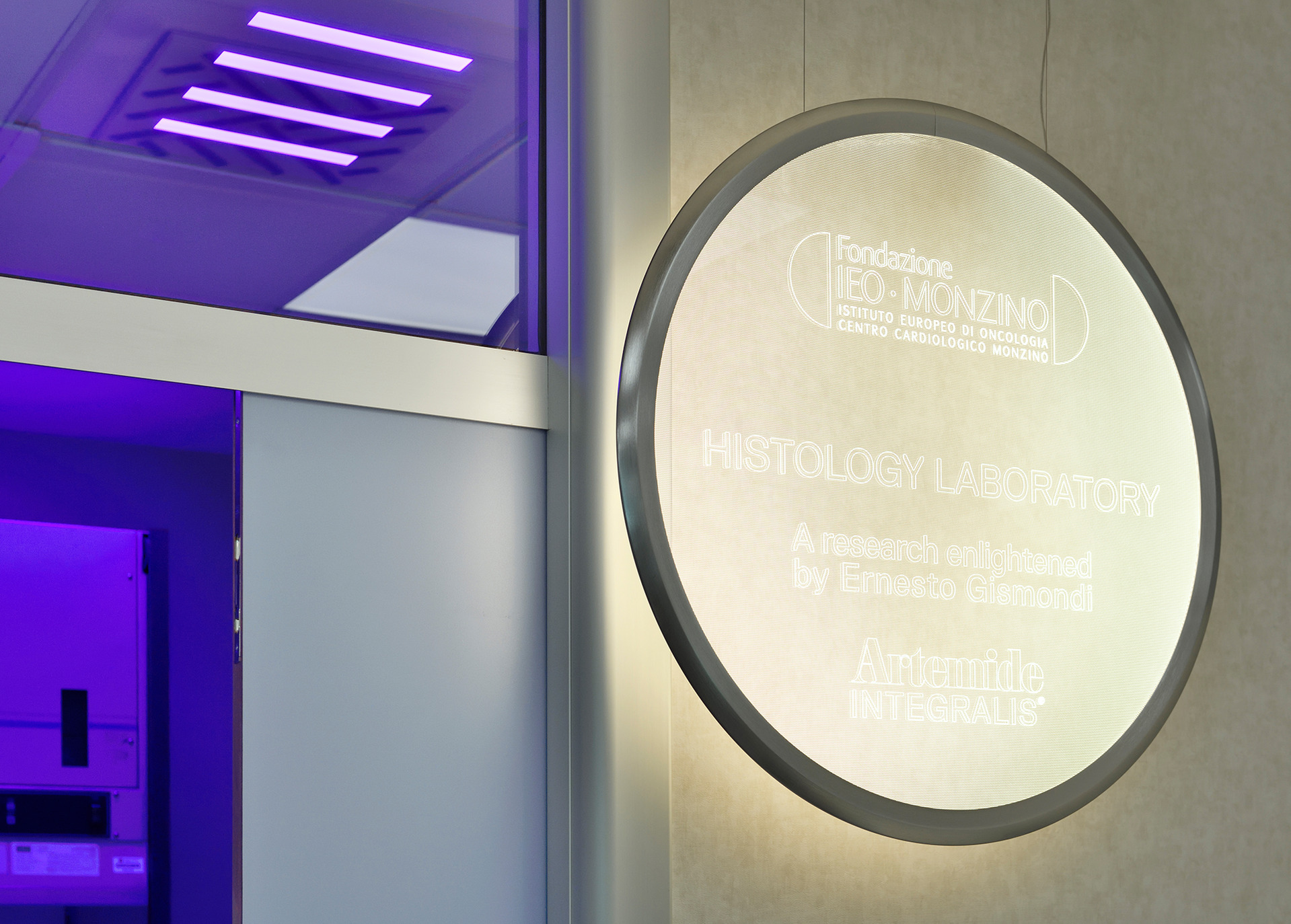 Illuminazione Artemide Integralis nel Laboratorio di Istologia della Fondazione IEO-Monzino, con luci a soffitto viola e un'insegna circolare luminosa.