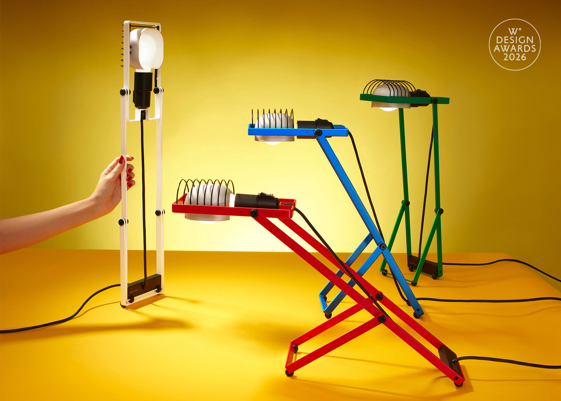 Quattro lampade Sintesi in bianco, rosso, blu e verde disposte su sfondo giallo con configurazioni diverse, fotografate da Pierpaolo Ferrari, con il logo di Wallpaper* Design Awards.