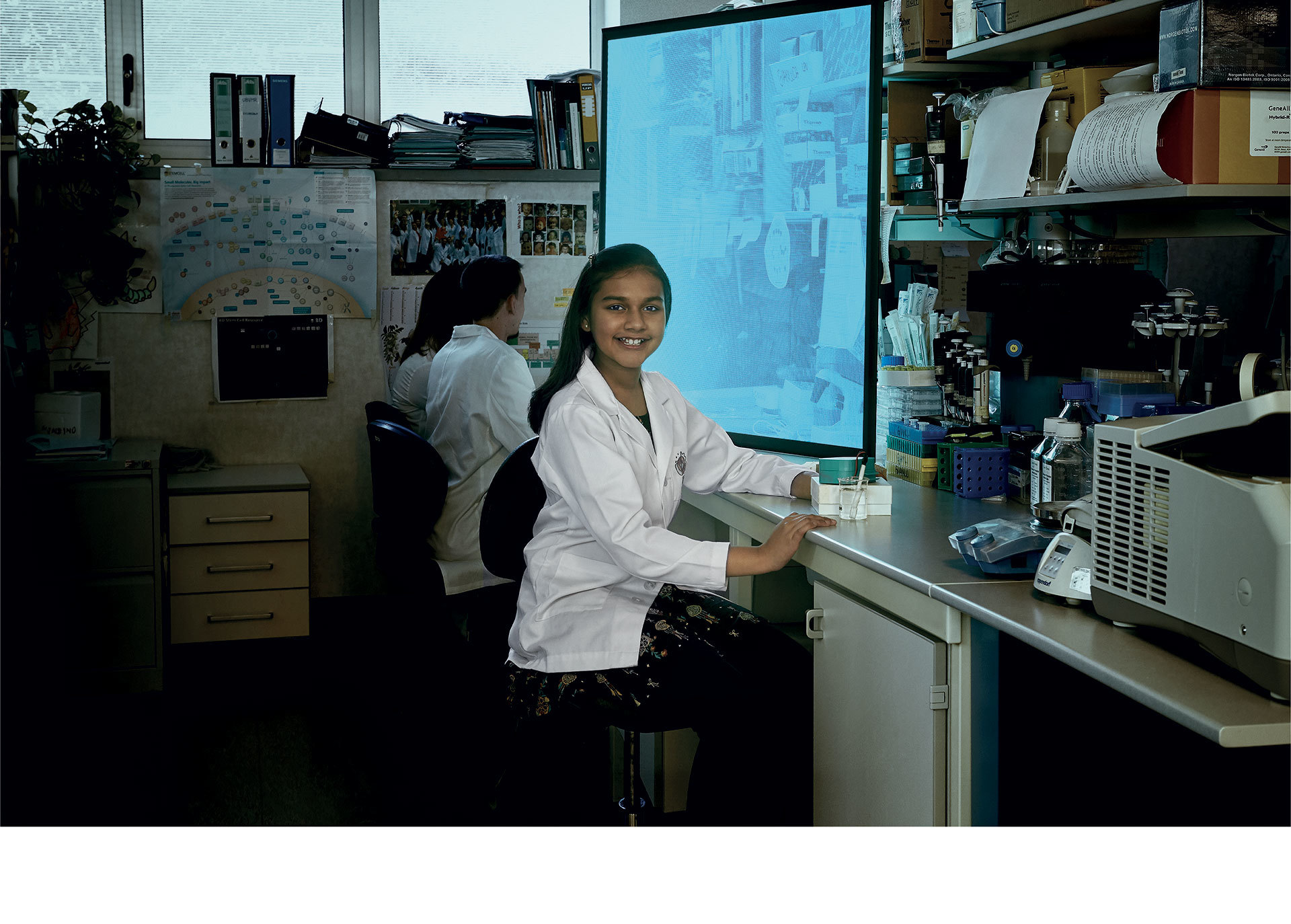 Due ricercatrici in camice bianco lavorano in un laboratorio, una sorridente verso la fotocamera, con attrezzature e un grande schermo sullo sfondo.