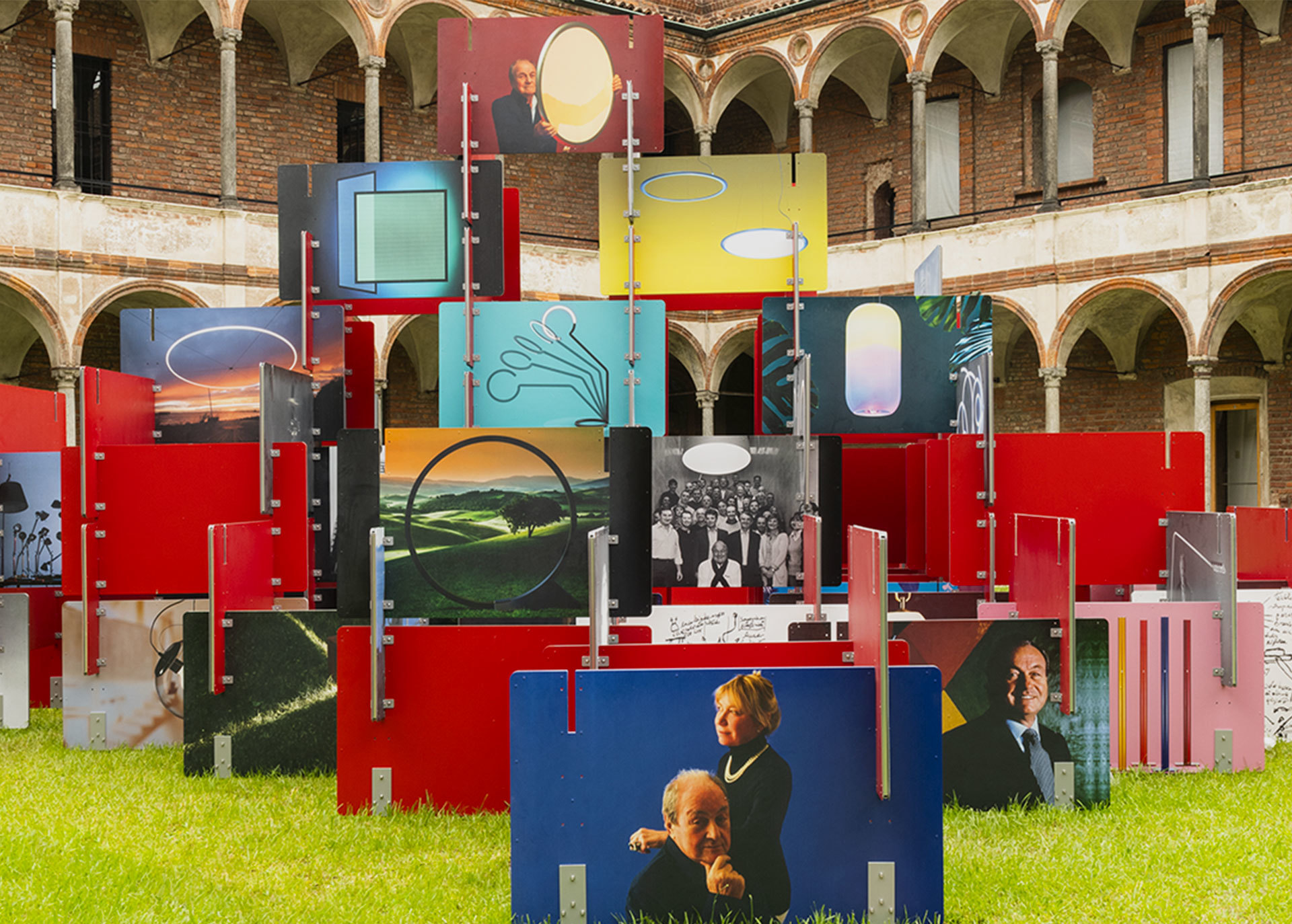 Installazione esterna colorata con pannelli rossi, fotografie e immagini di lampade Artemide in un cortile storico porticato.