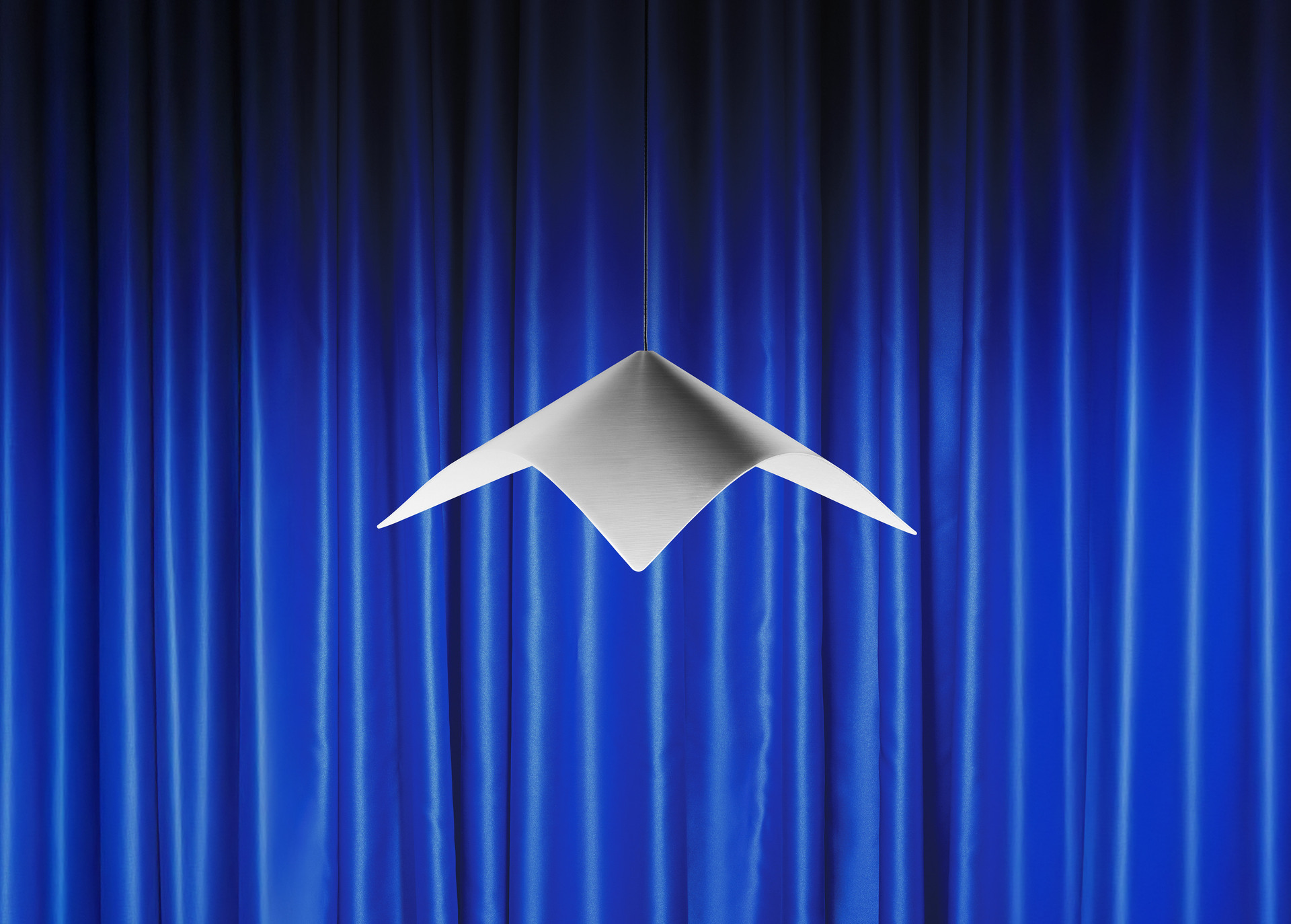 Lampada a sospensione Iperbole davanti a tende da teatro blu.
