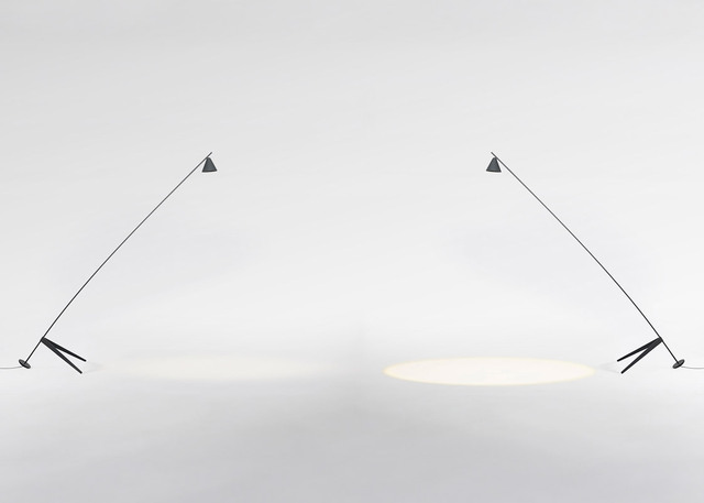 Due lampade da terra minimaliste scure con bracci lunghi inclinati e paralumi conici, che proiettano fasci di luce concentrati su una superficie bianca.