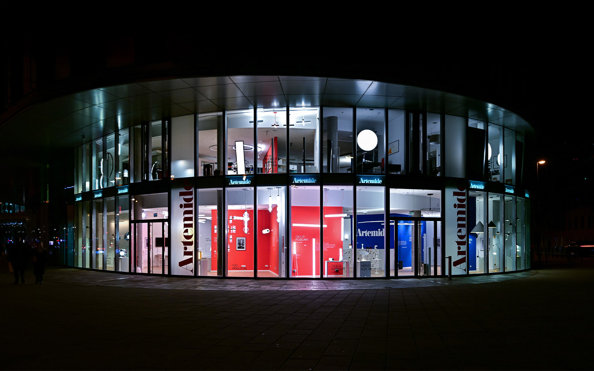 Esterno del flagship store Artemide di notte, facciata curva in vetro illuminata con aree espositive rosse e blu che mostrano le collezioni di illuminazione