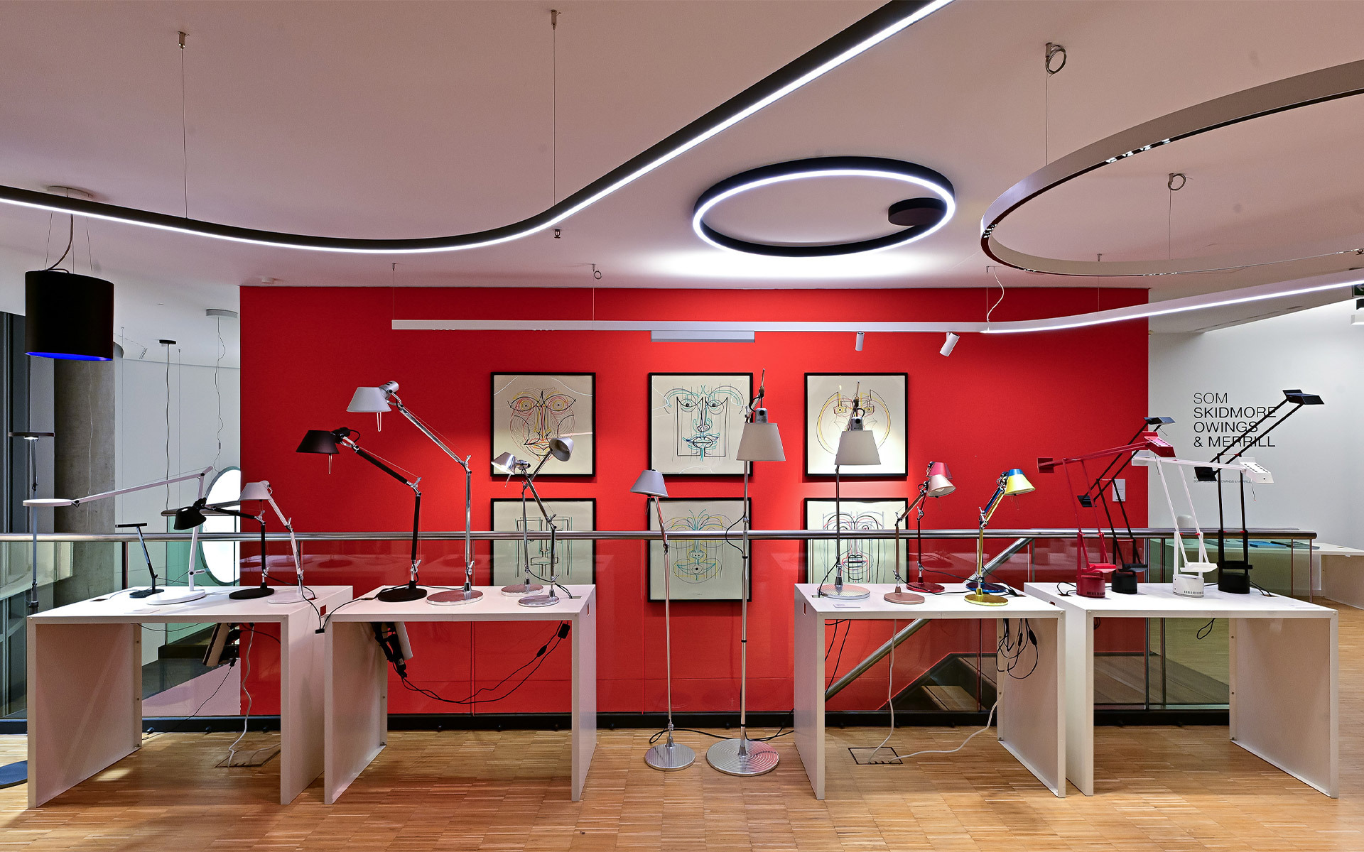 Showroom Artemide con lampade da tavolo e da terra Tolomeo su tavoli bianchi davanti a una parete rossa, con luci circolari LED a soffitto.