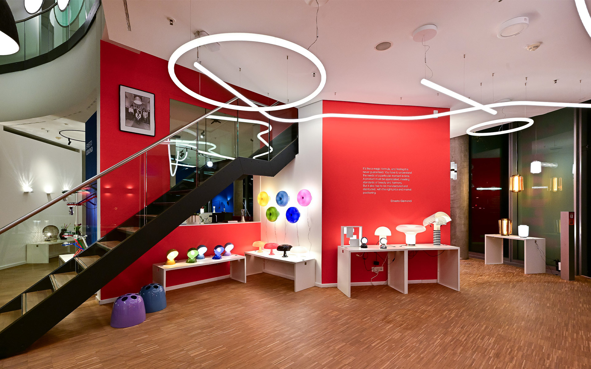Showroom Artemide con lampade da tavolo colorate, luci a sospensione circolari e ondulate su pareti rosse, scala e lampade su tavoli bianchi.