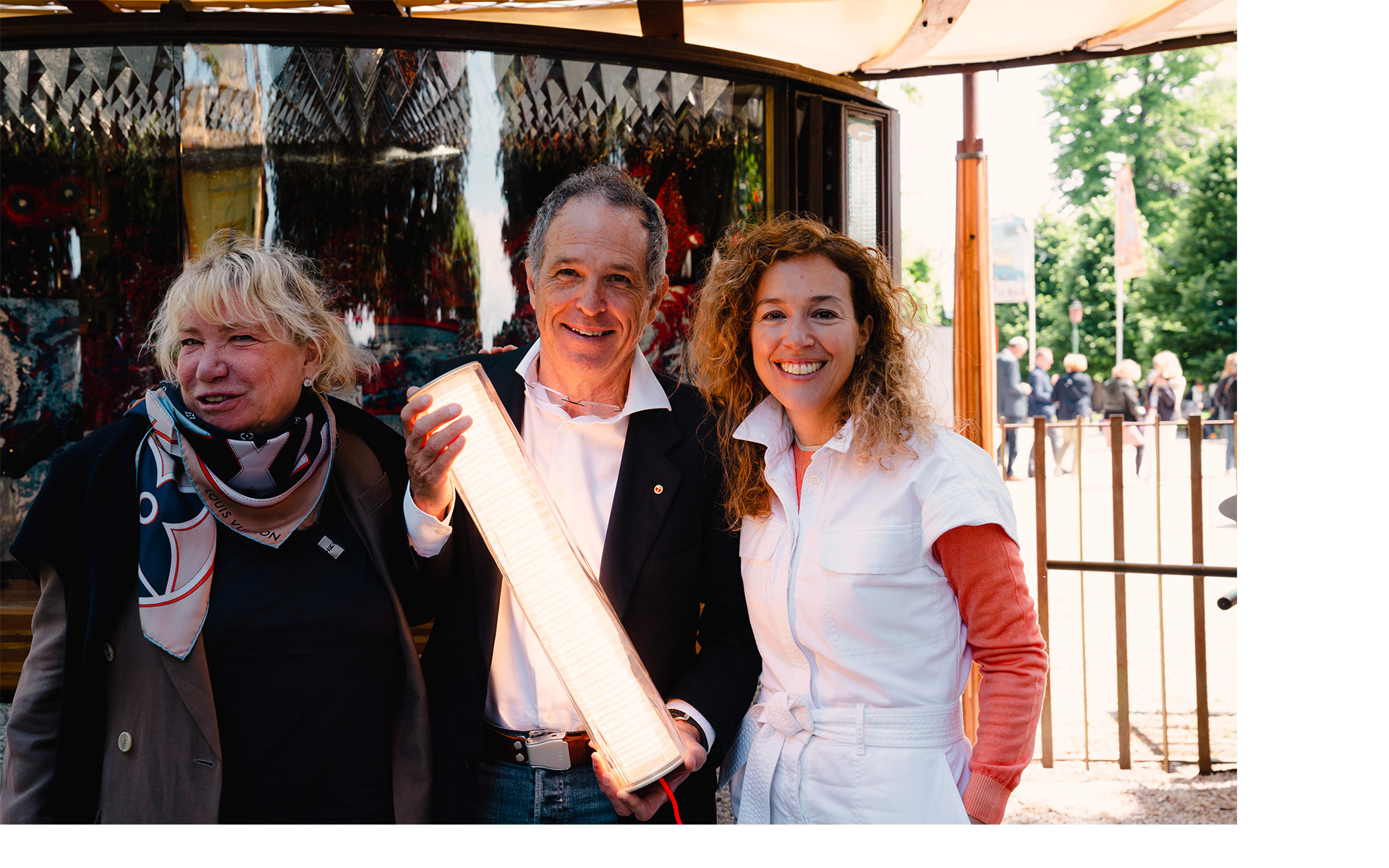Tre persone sorridenti all'aperto, la persona al centro tiene una lampada LED tubolare Artemide illuminata a un evento.