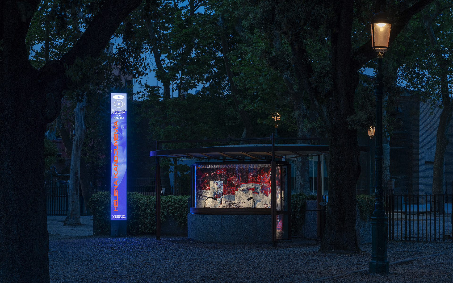 Chiosco esterno illuminato e insegna verticale luminosa al crepuscolo, circondati da alberi, con un lampione classico che diffonde luce calda