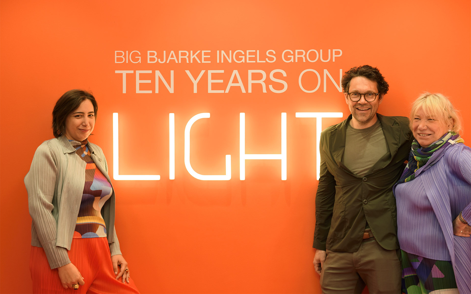 Tre persone in posa davanti a una parete arancione con l'insegna luminosa 'BIG Bjarke Ingels Group Ten Years on Light'.