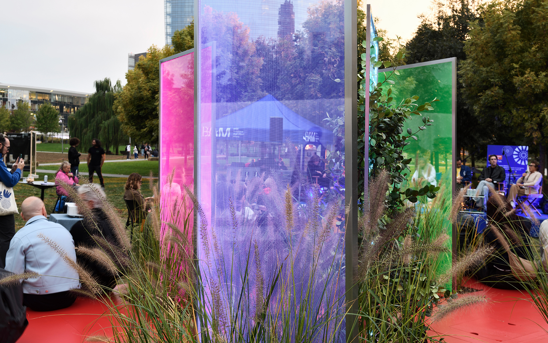 Outdoor installation with tall colorful transparent panels in pink, blue, and green, surrounded by ornamental grasses and people at dusk.