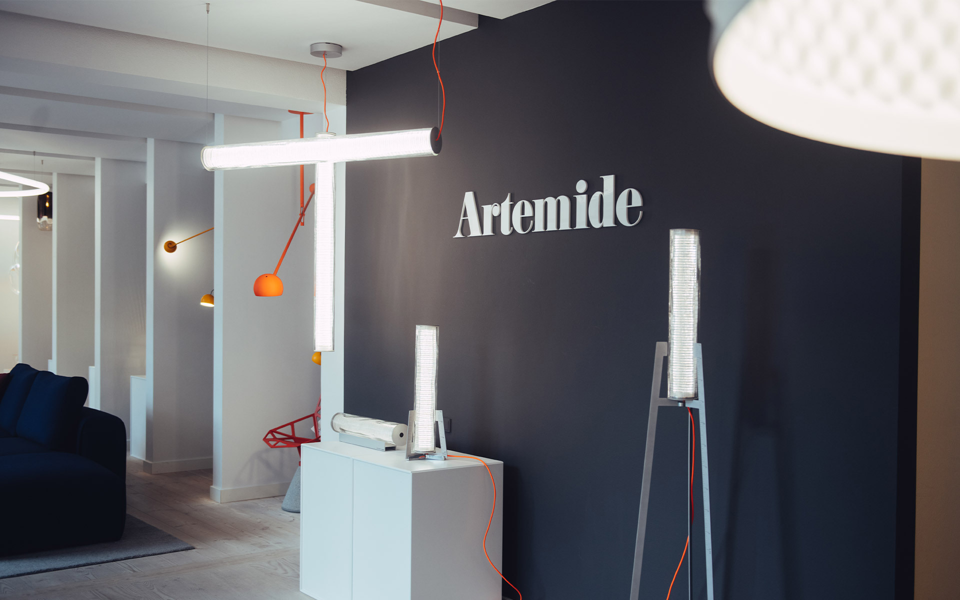 Showroom Artemide con diverse luminarie: sospensione tubolare LED orizzontale, lampade da parete arancioni e lampade da tavolo su piedistalli bianchi.