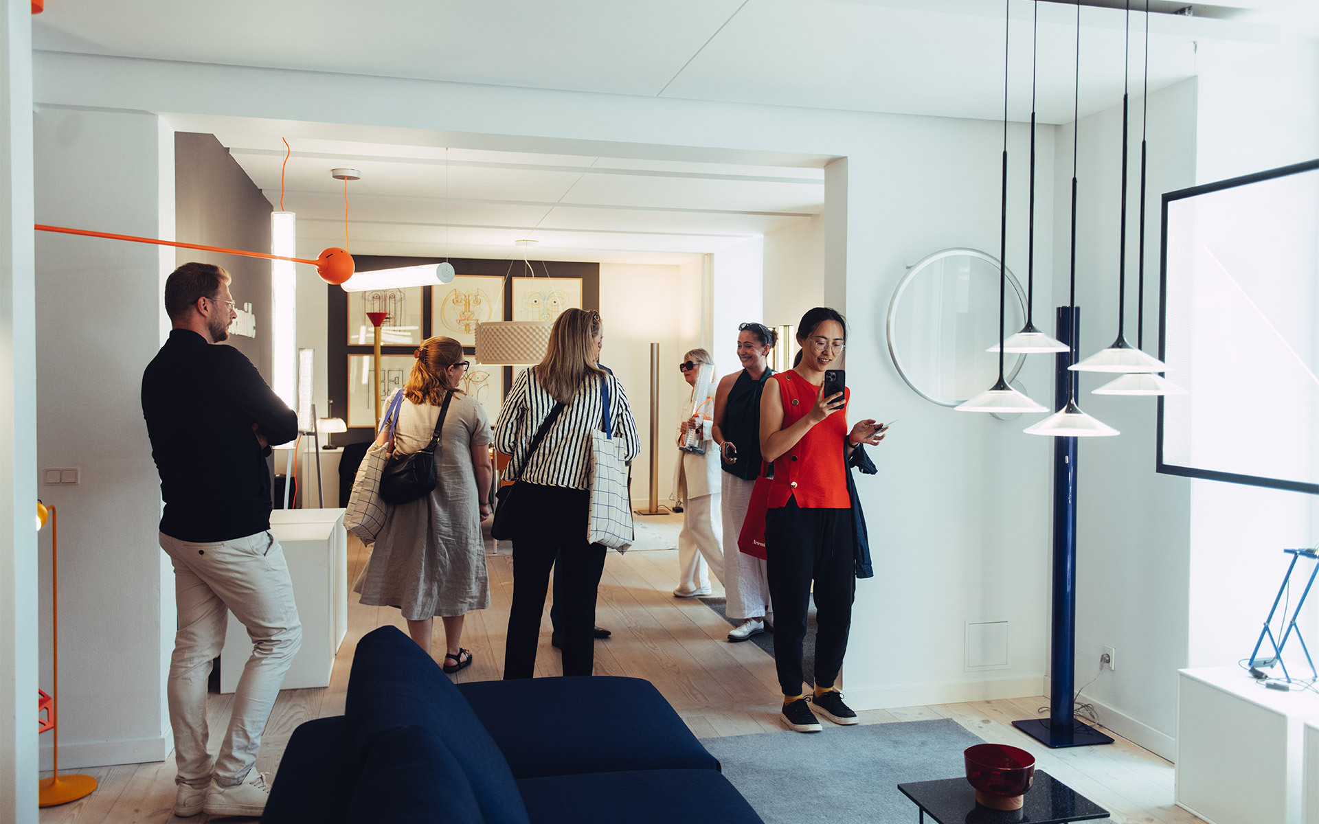Visitatori che esplorano uno showroom Artemide con lampade a sospensione e da terra in tonalità bianco, arancione e blu.