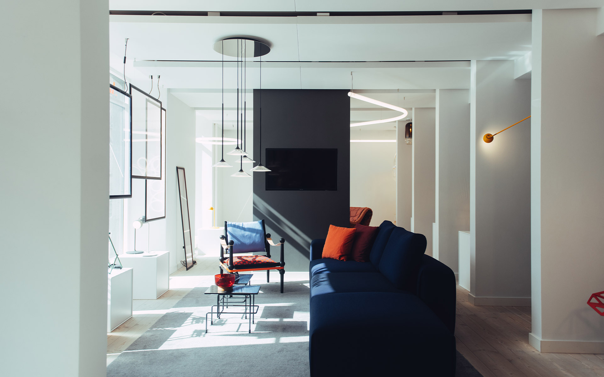 Soggiorno moderno con sospensione LED circolare Artemide, sottili pendenti neri e lampada da parete dorata sopra un divano blu navy con cuscini rossi
