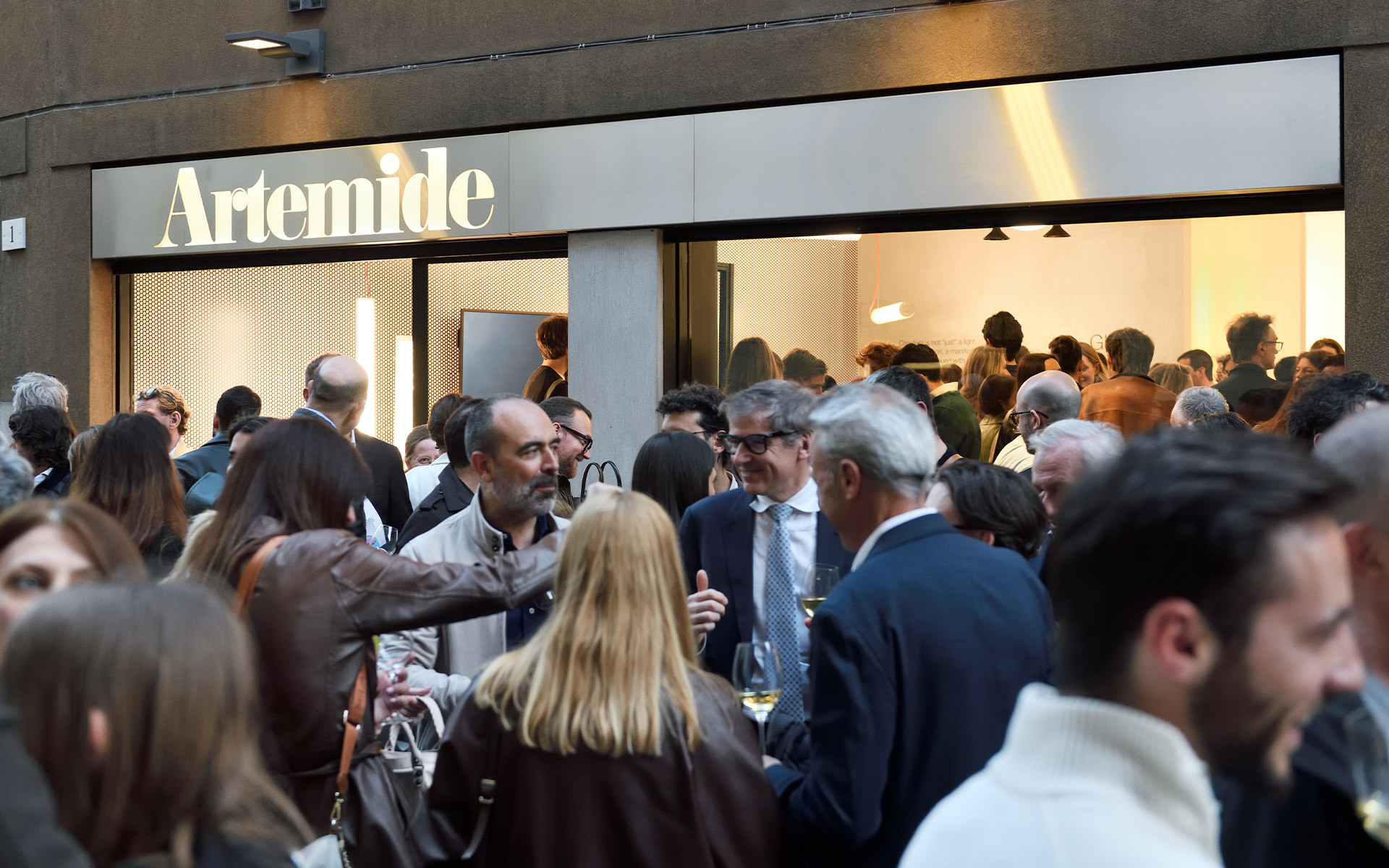 Folla di ospiti riunita fuori da un negozio Artemide durante un evento pubblico o un'inaugurazione, con l'insegna luminosa del marchio sopra.