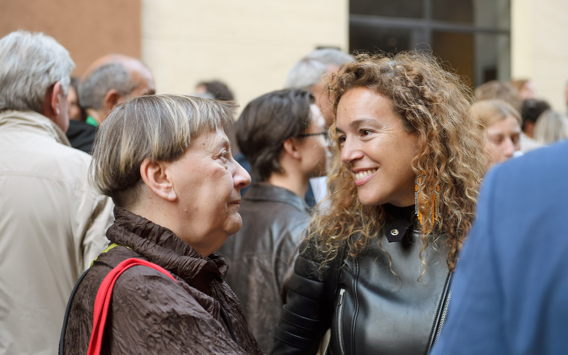 Due donne in conversazione a un evento sociale, una più anziana con capelli grigi corti, una più giovane con capelli ricci, sorridente.