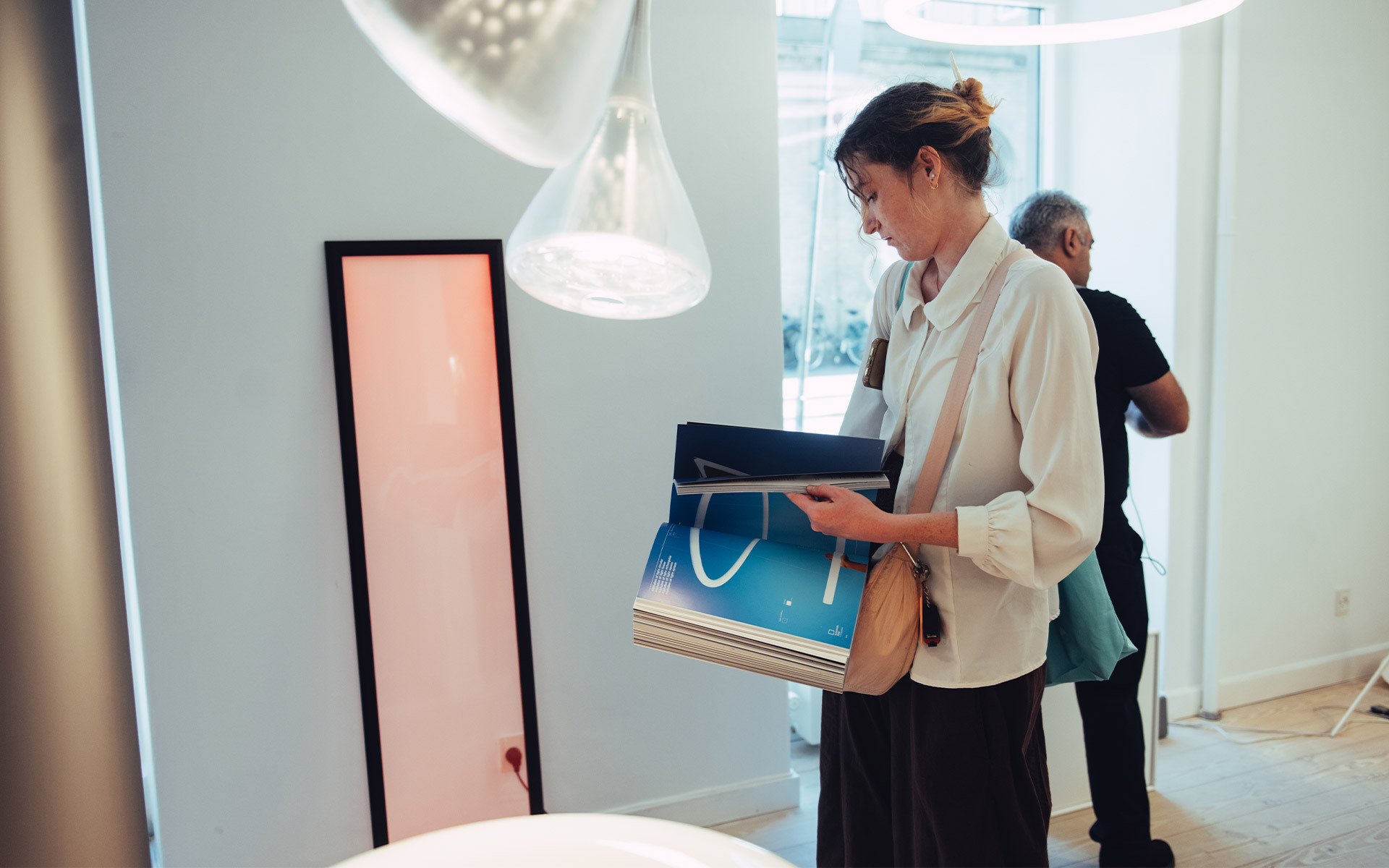 Donna che esplora un moderno showroom di illuminazione, con una borsa della spesa, una lampada a sospensione bianca e una lampada da terra rosa vicino a lei.