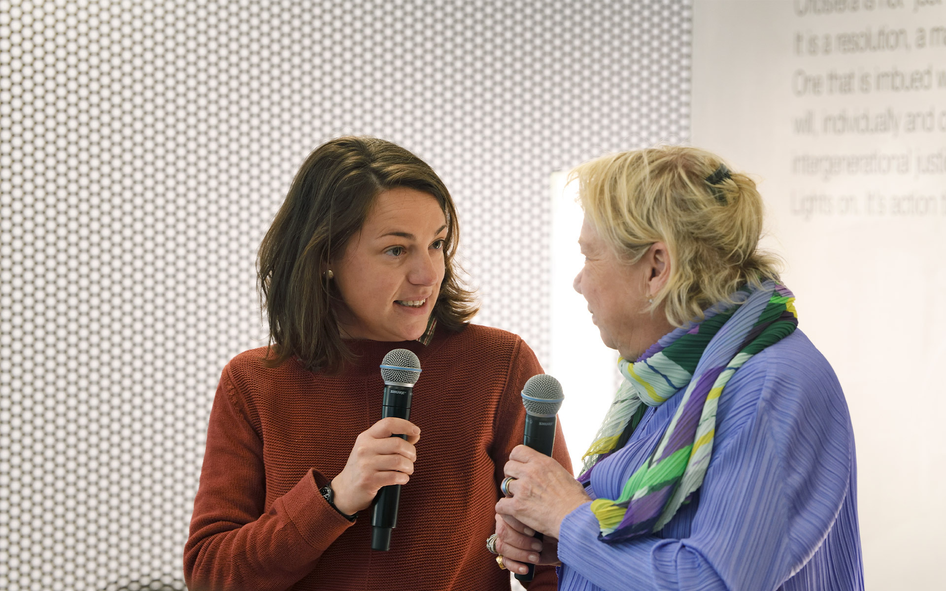 Due donne con microfoni in conversazione a un evento Artemide, con pannello a pois e testo espositivo sullo sfondo.