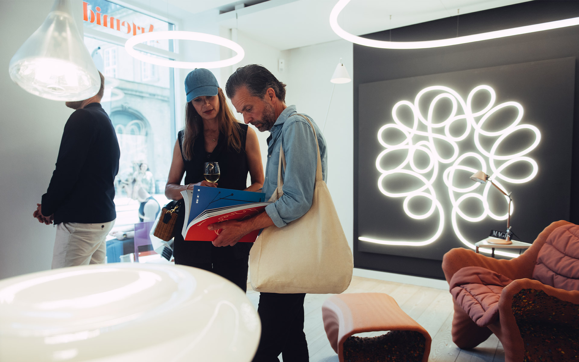 Visitatori sfogliano cataloghi in uno showroom con lampada da parete Artemide a LED a forma di albero e lampade a sospensione circolari.