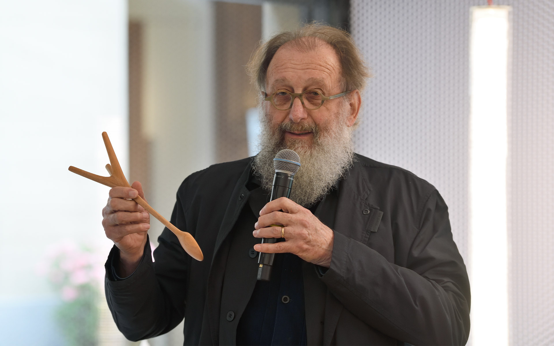 Uomo con barba in giacca scura parla al microfono tenendo un cucchiaio di legno e un oggetto a forma di forchetta durante una presentazione