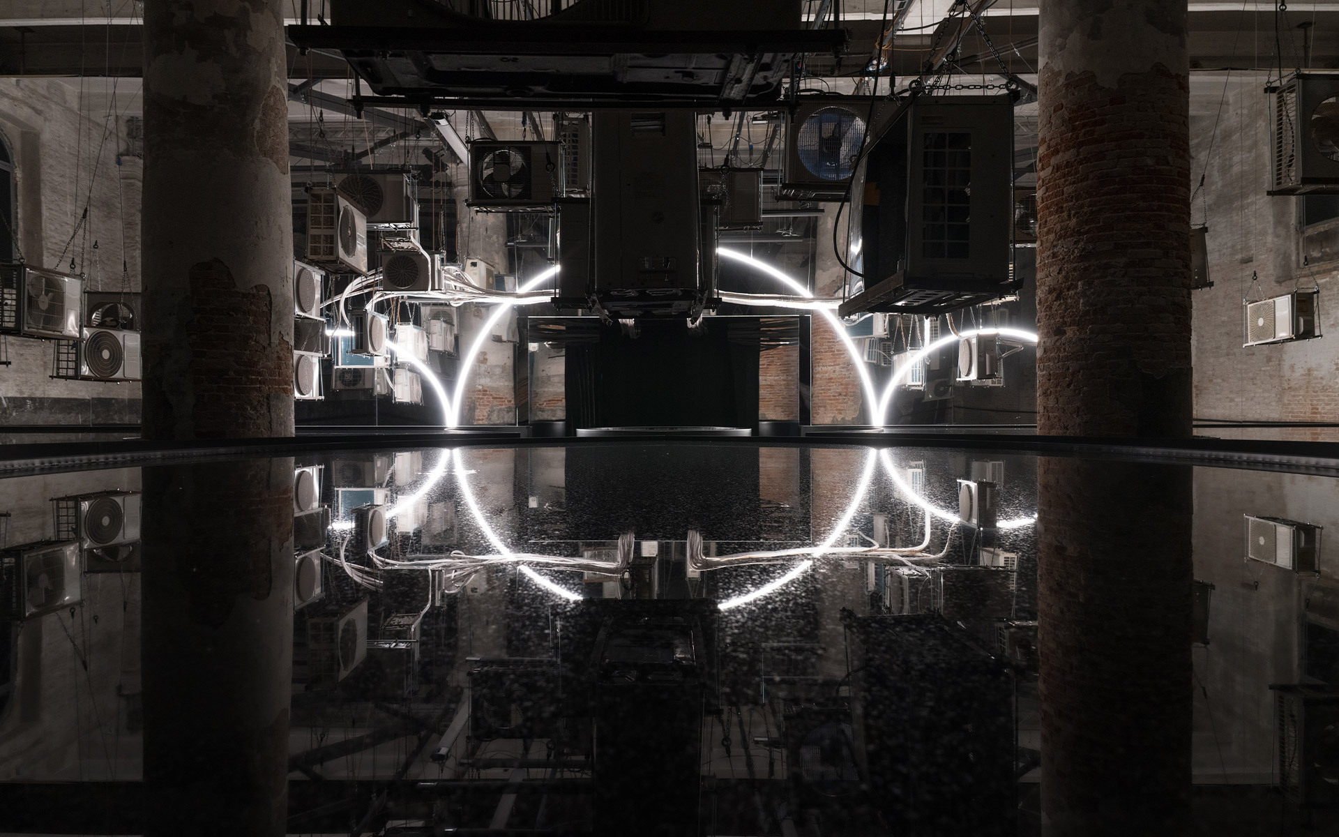 Installazione luminosa tubolare bianca a semicerchio, riflessa in una vasca scura in uno spazio industriale con colonne in mattoni.