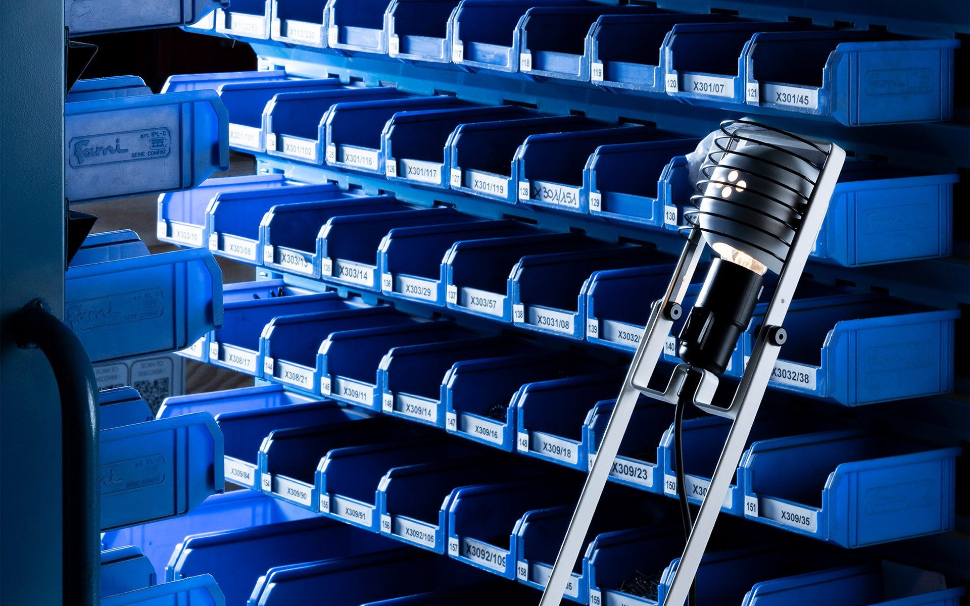 Lampada Artemide con testa cilindrica nera su una scala, che illumina scaffali industriali blu con contenitori etichettati