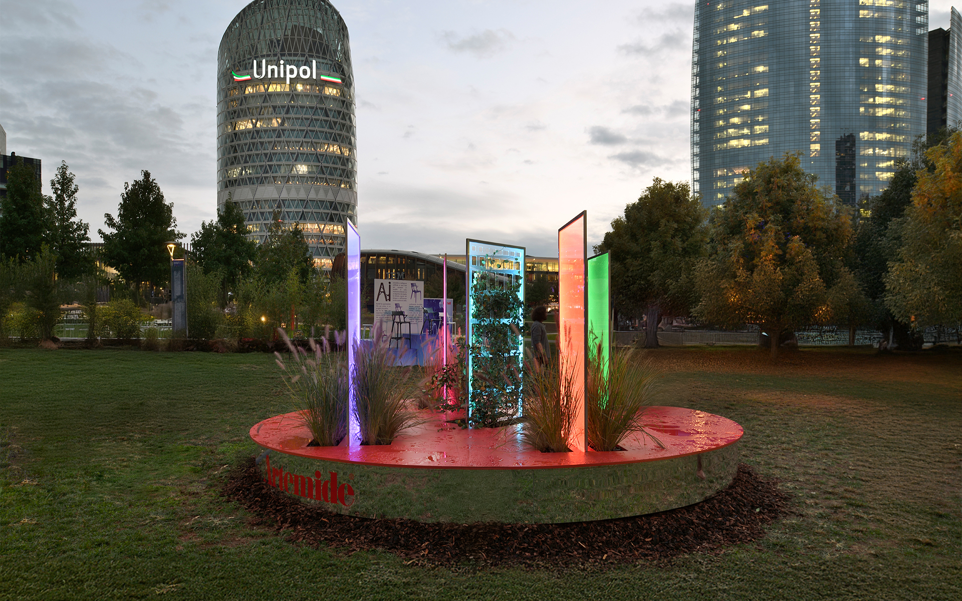 Colorful illuminated vertical panels installation on a circular platform in an urban park at dusk, with the Unipol tower in the background