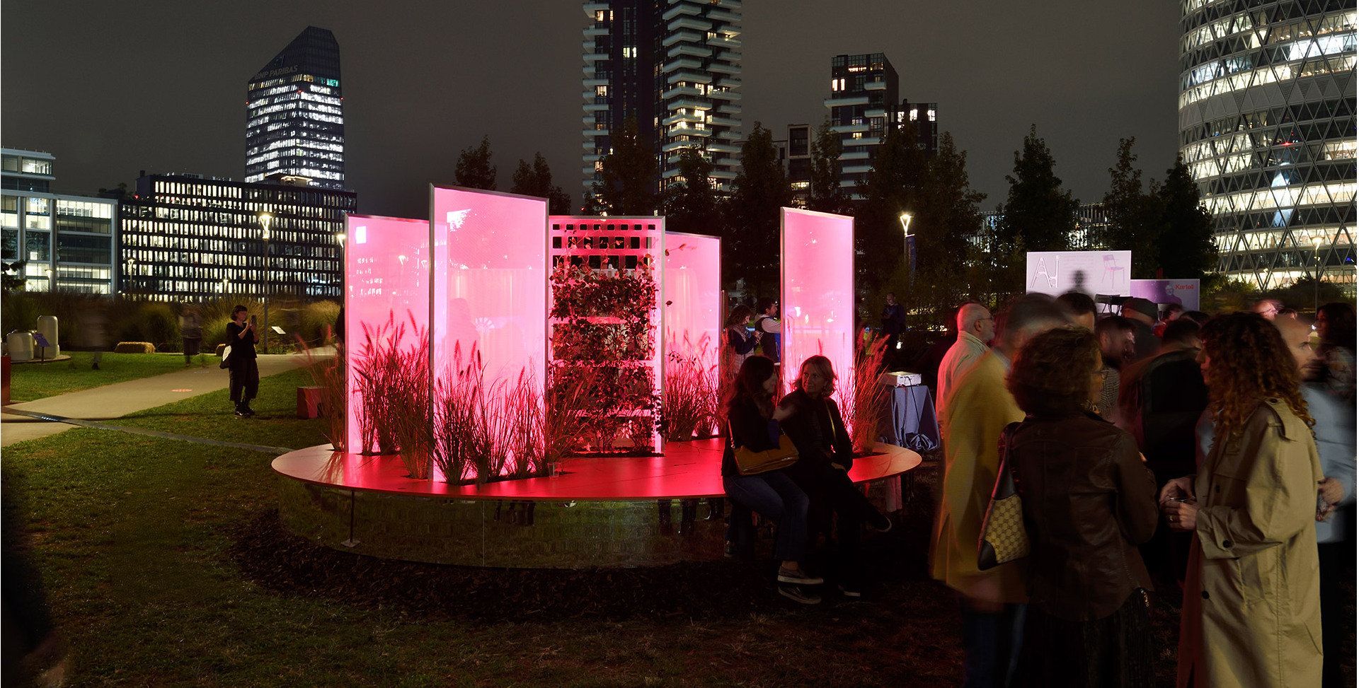 Outdoor installation with red-lit vertical panels and plants on a circular platform, surrounded by people at night with city skyline.