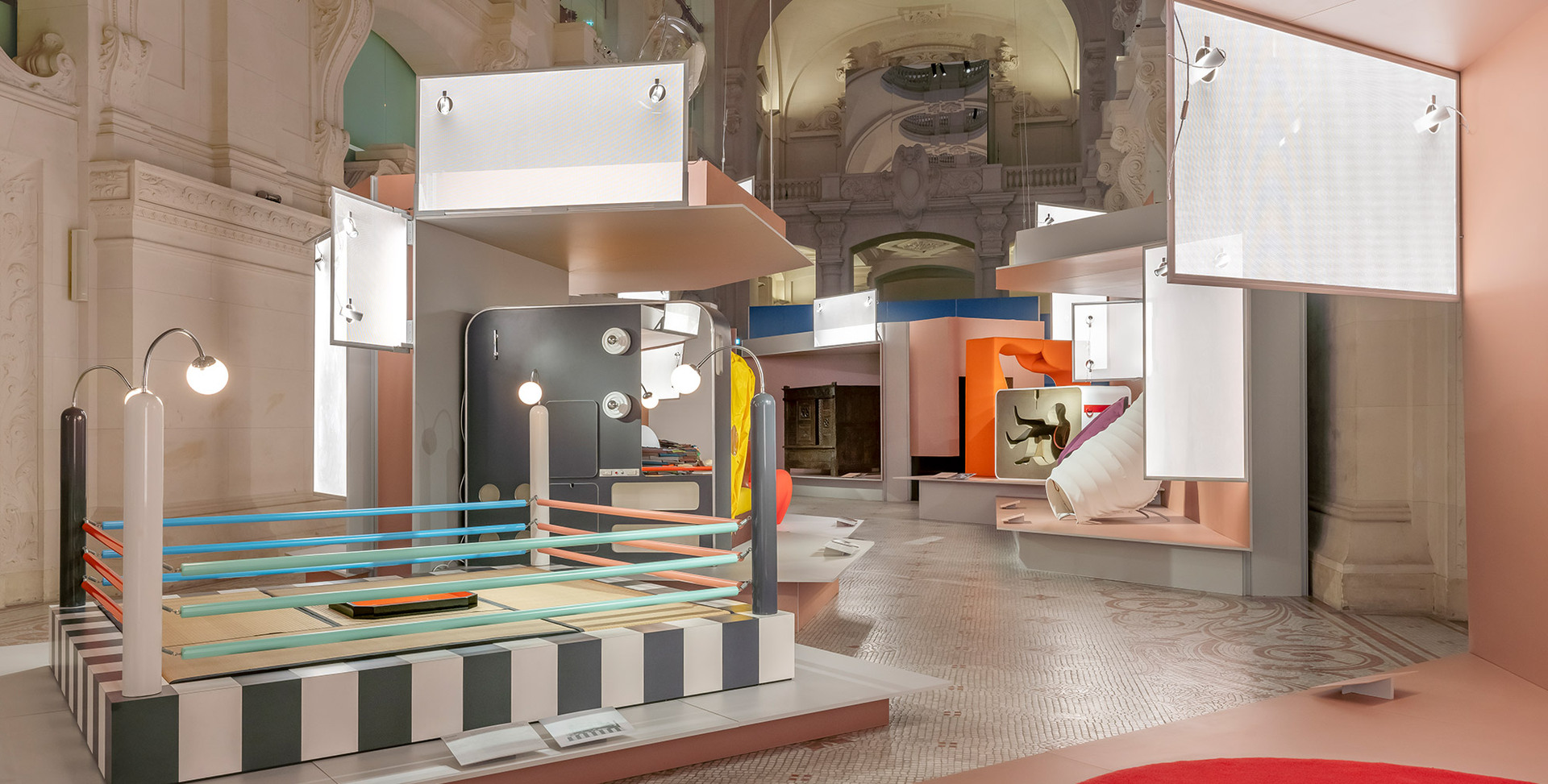 Installazione espositiva colorata in una sala storica con volte, strutture geometriche, pannelli sospesi e lampade Artemide.