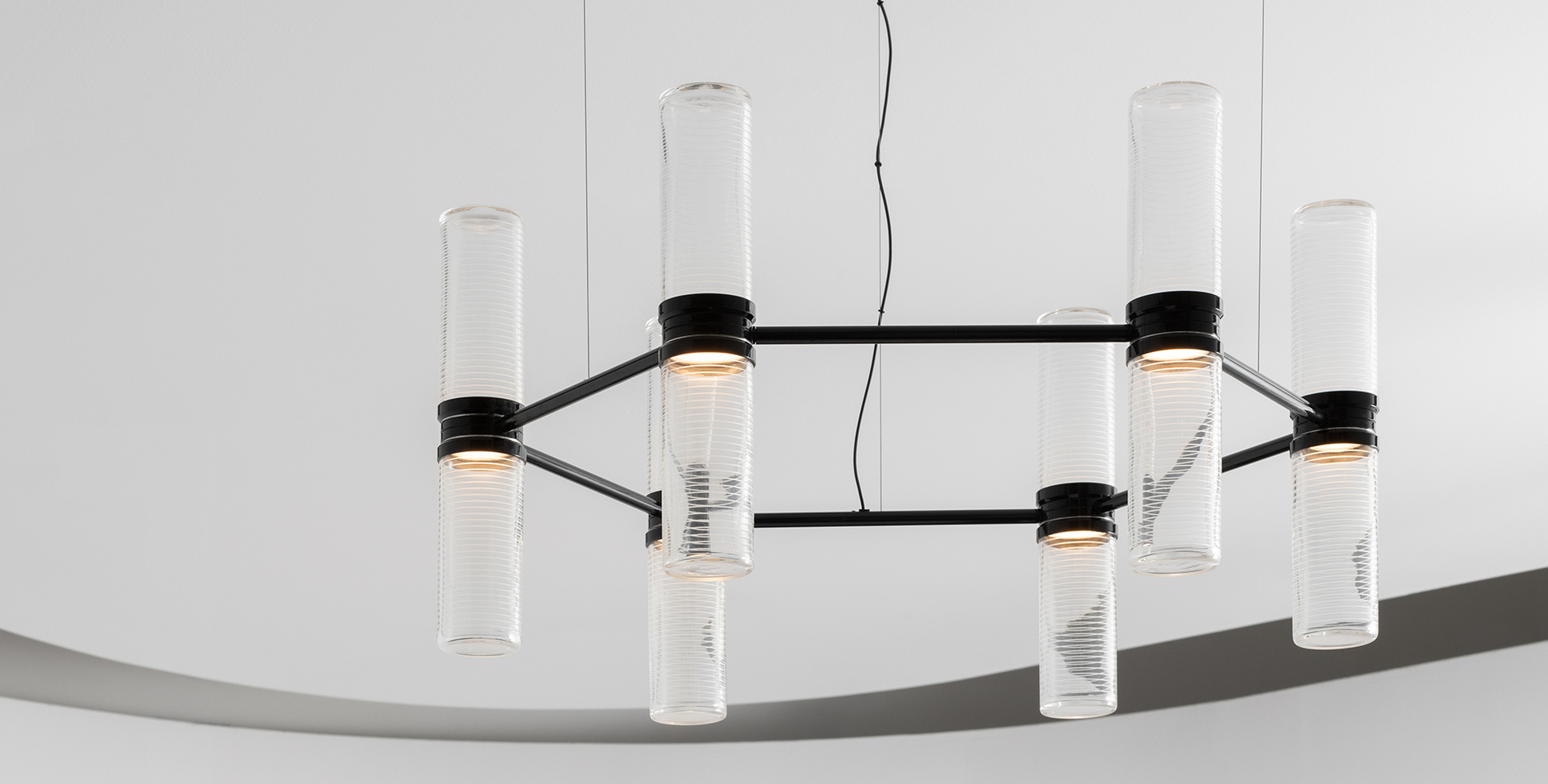 Lampadario a sospensione con struttura rettangolare nera che sostiene otto tubi cilindrici verticali in vetro trasparente con sorgenti LED calde