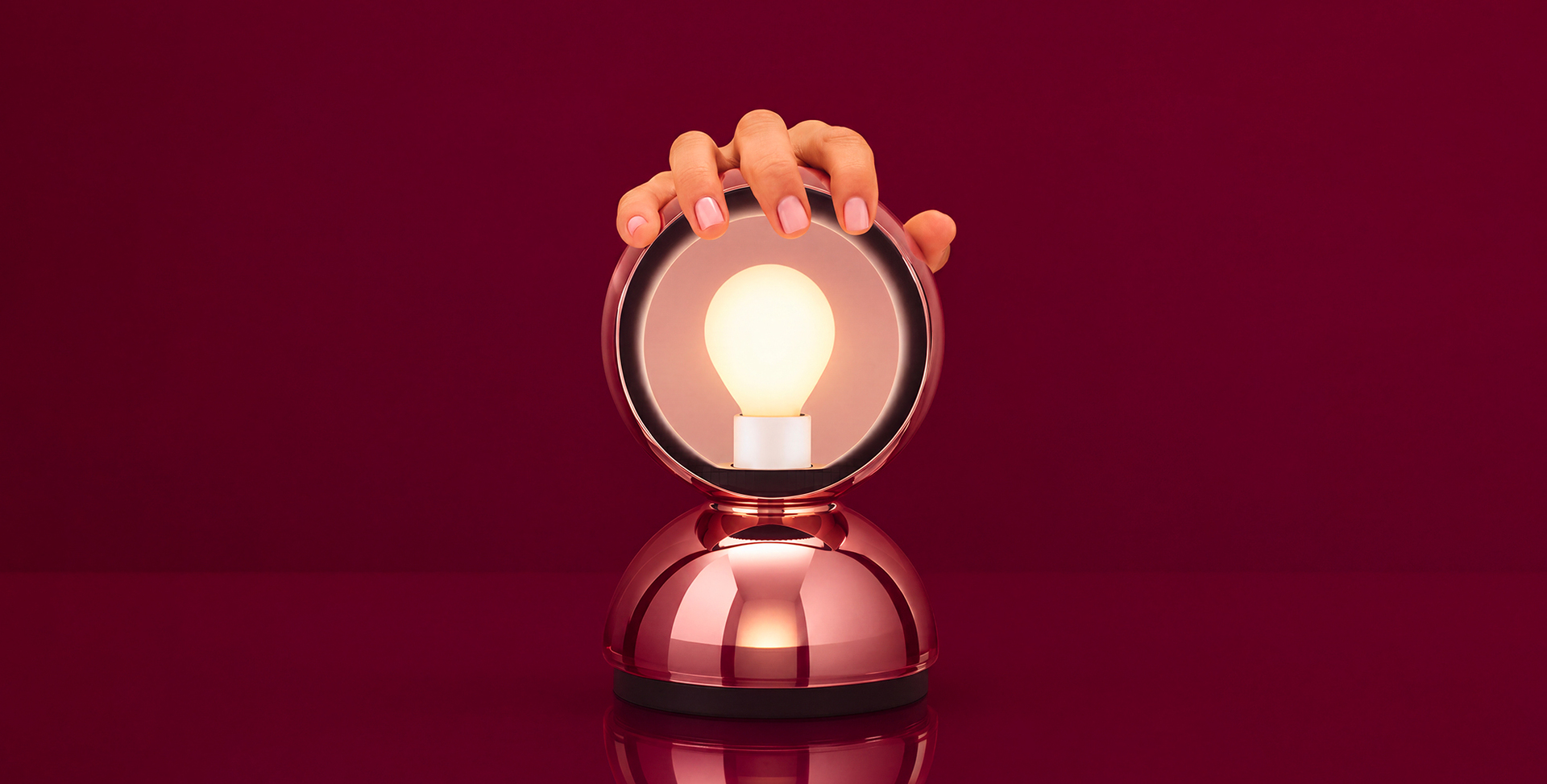 Hand touching a glowing bulb inside a rose gold metallic spherical table lamp with a rounded chrome base.
