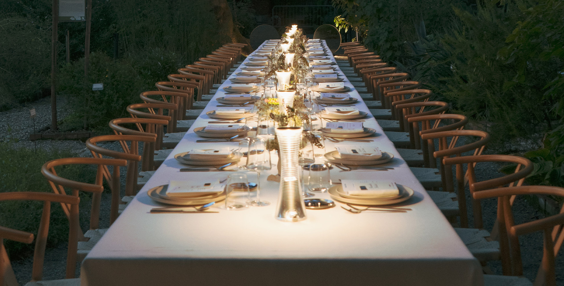 Lunga tavolata all'aperto in un giardino serale, con sedie in legno, piatti eleganti e lampade "Come together" appoggiate sul tavolo.