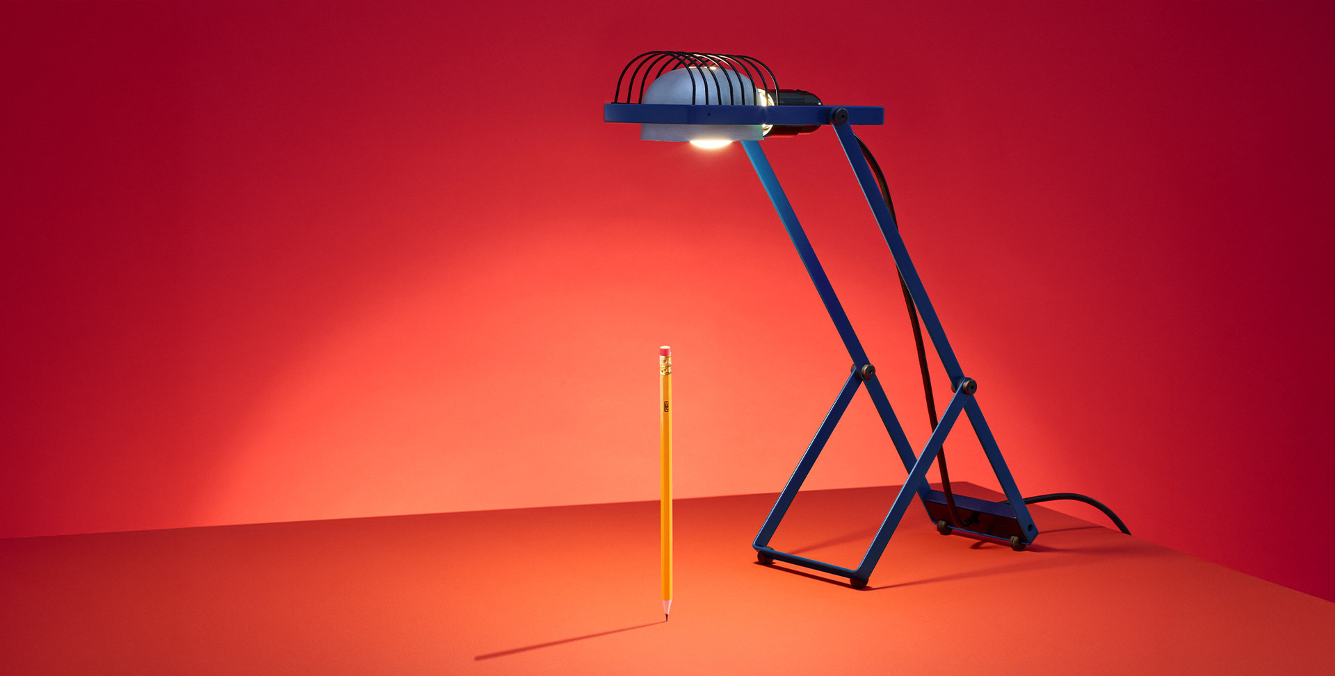 Lampada Sintesi blu che illumina una matita verticale su sfondo rosso, fotografata da Pierpaolo Ferrari.