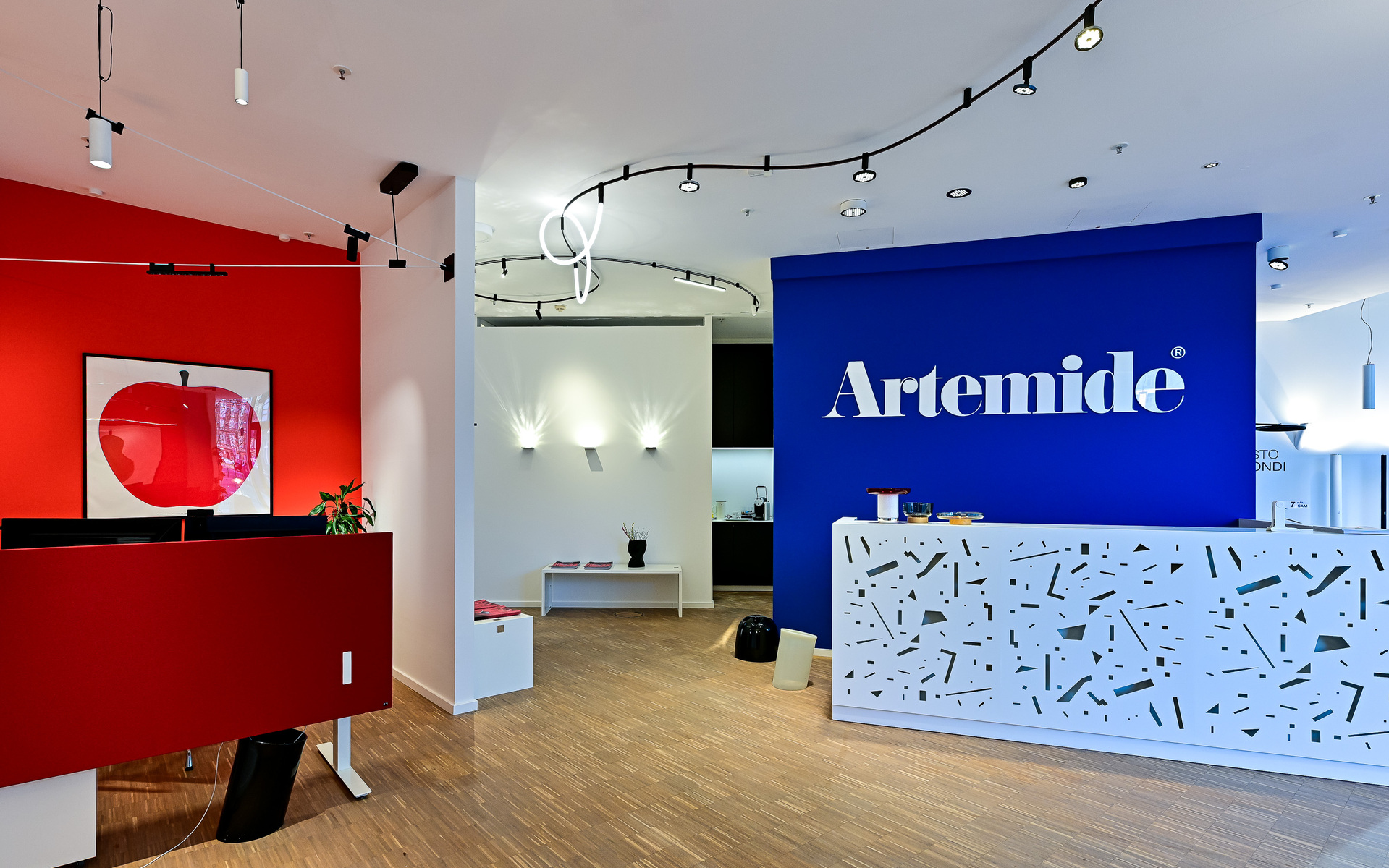 Ingresso dello showroom Artemide con reception blu brandizzata, parete rossa, faretti su binario a soffitto e applique a parete.