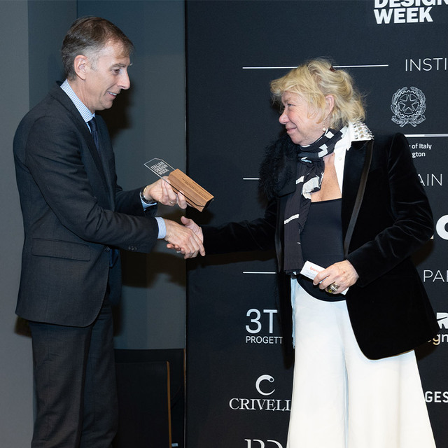 Due persone si stringono la mano a una cerimonia di premiazione del Design Week, uomo in abito scuro consegna un trofeo in legno a una donna in giacca nera.