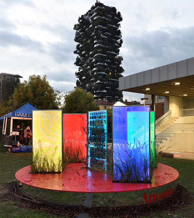 Artemide installation: four illuminated colored panels (yellow, red, blue, cyan) with grass motifs on a red platform, Bosco Verticale in background, Milan