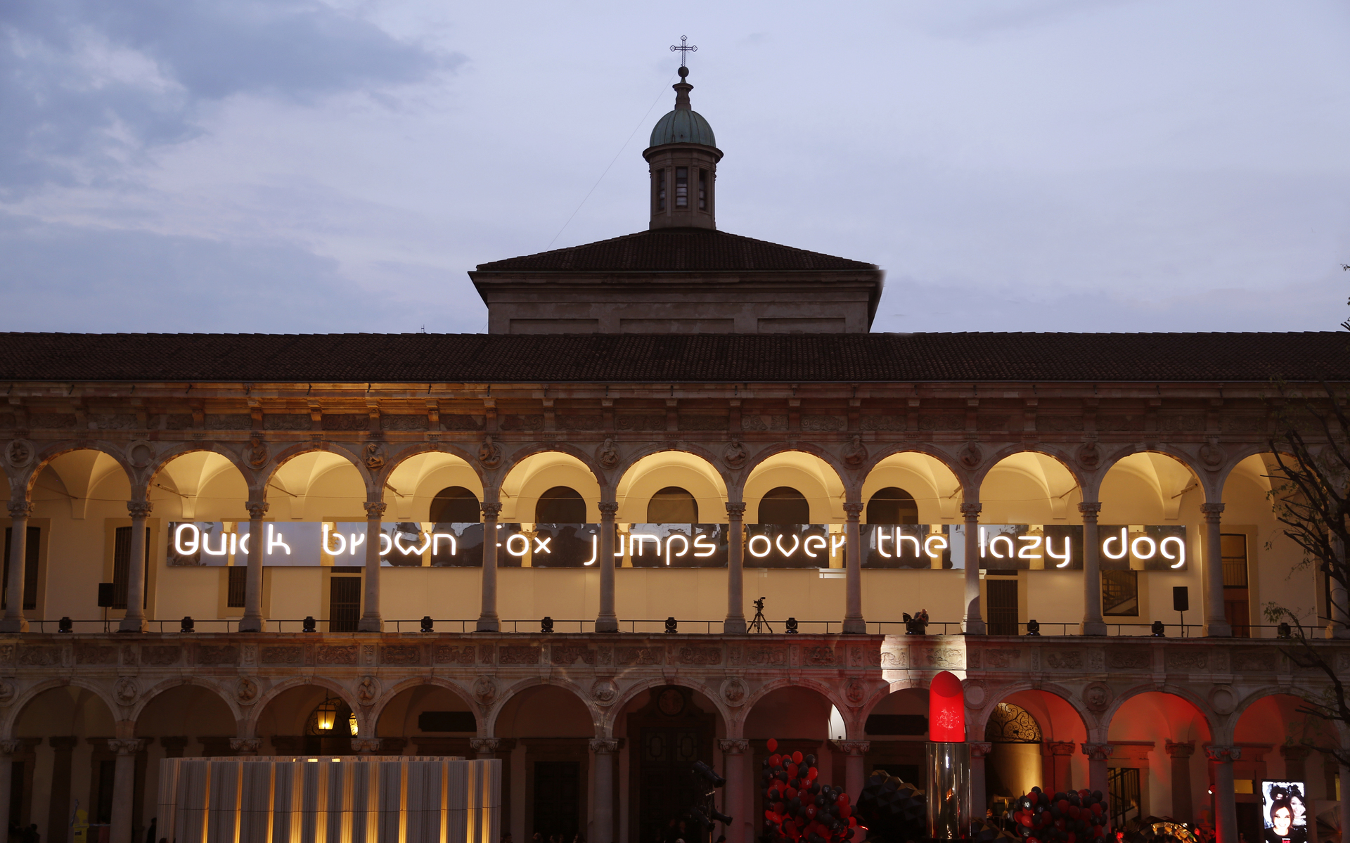 Testo illuminato 'Quick brown fox jumps over the lazy dog' proiettato sul porticato di un edificio classico italiano al crepuscolo