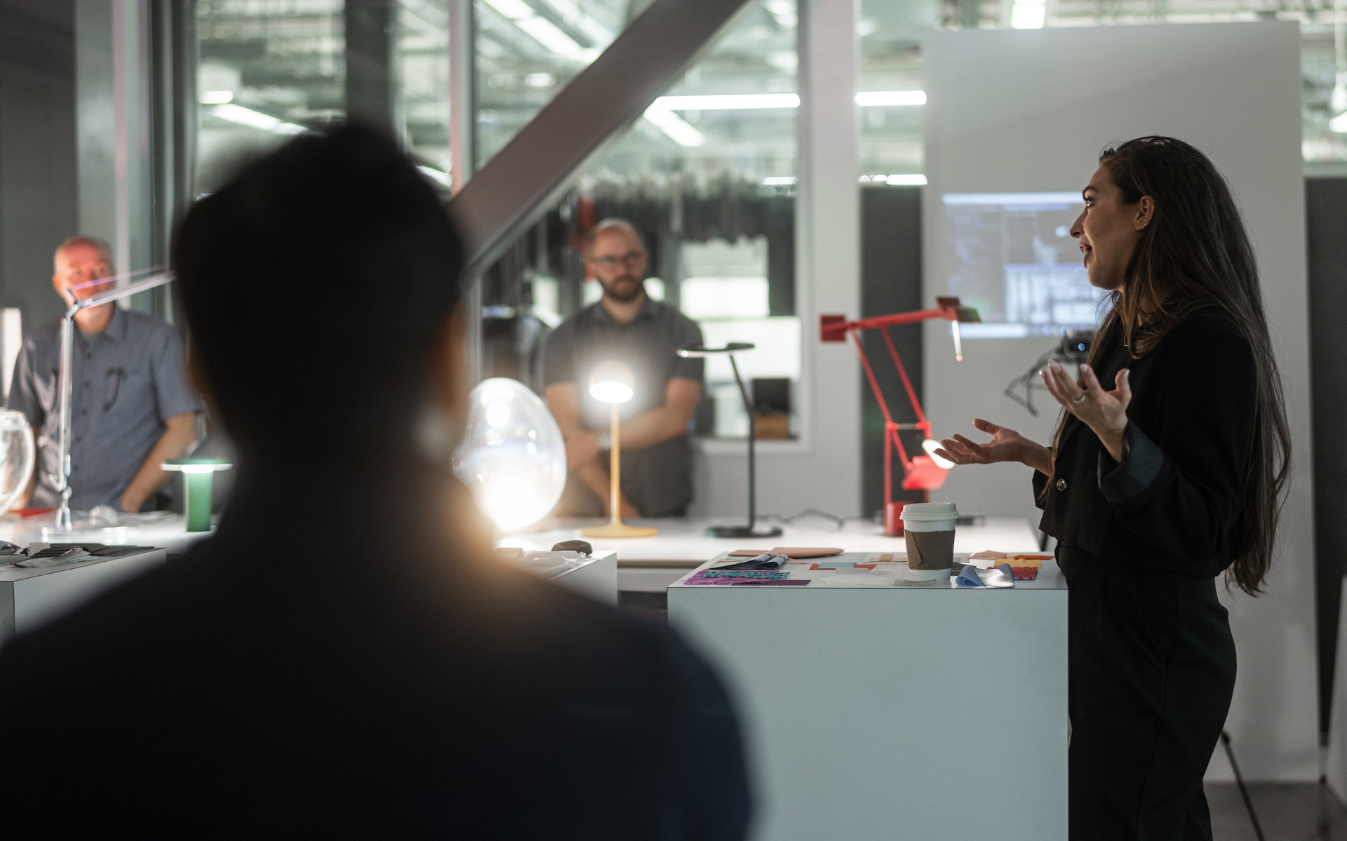 Una presentatrice parla ai colleghi in uno studio di design con lampade da tavolo Artemide, tra cui una Tolomeo rossa e una lampada a globo luminosa sulla scrivania.