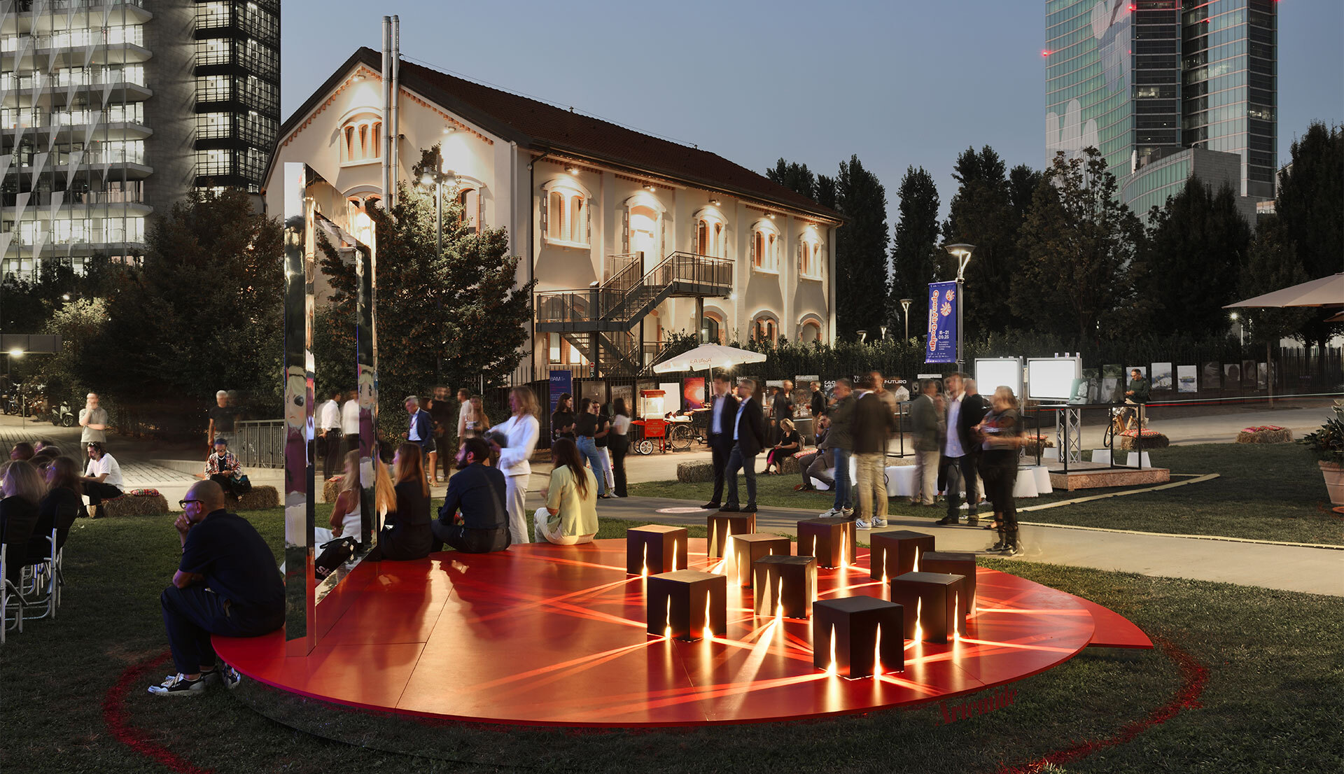 Evento serale all'aperto con persone riunite attorno a una piattaforma circolare rossa luminosa con sgabelli in legno, illuminata dal basso, contesto urbano.