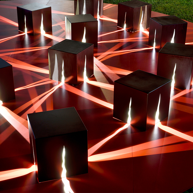 Cubi luminosi neri su pedana rossa con fasci di luce che si incrociano creando geometrie.