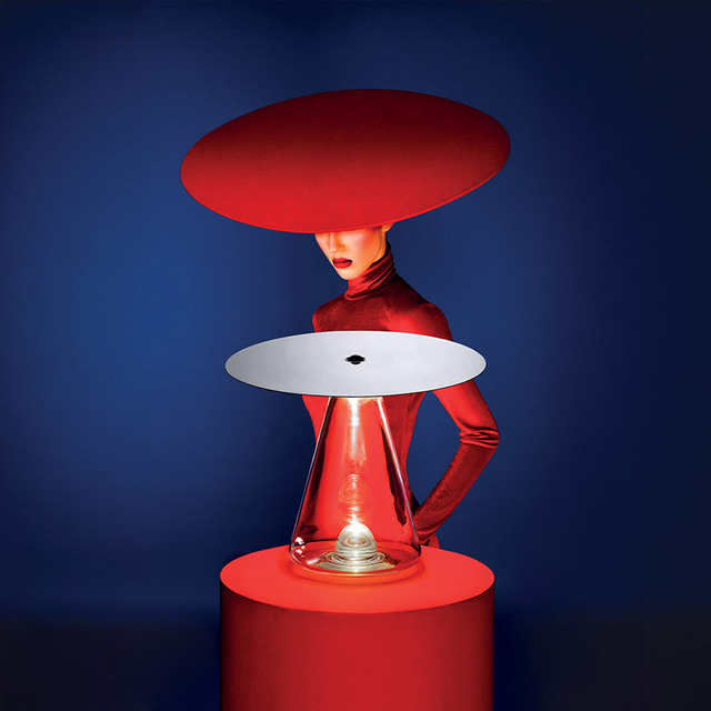 Lampada da tavolo Artemide con base conica trasparente e paralume a disco piatto su piedistallo rosso, abbinata a modella in rosso con cappello a tesa larga