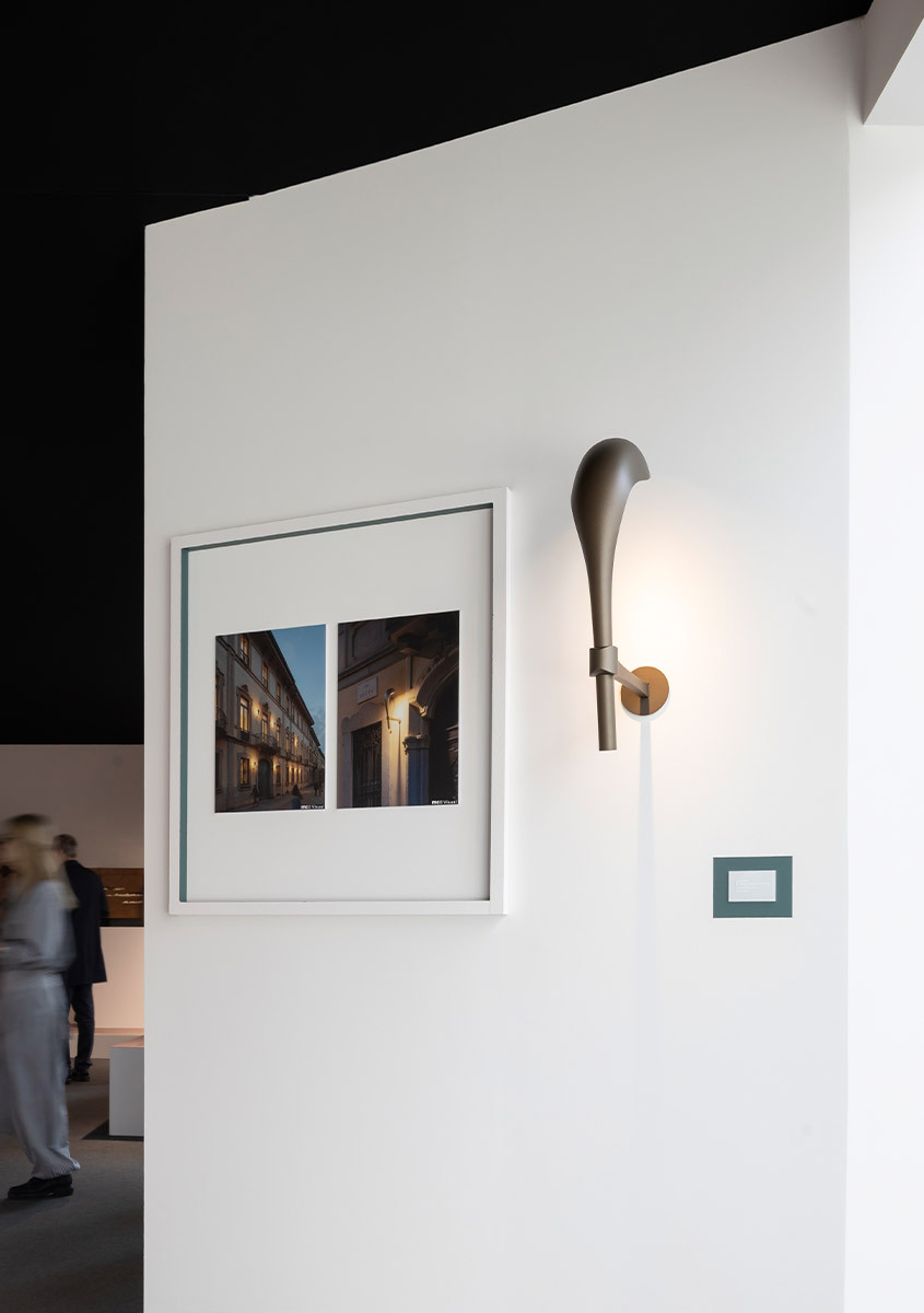 Applique Artemide con paralume conico allungato scuro e supporto in ottone, esposta su parete bianca accanto a fotografia architettonica incorniciata.