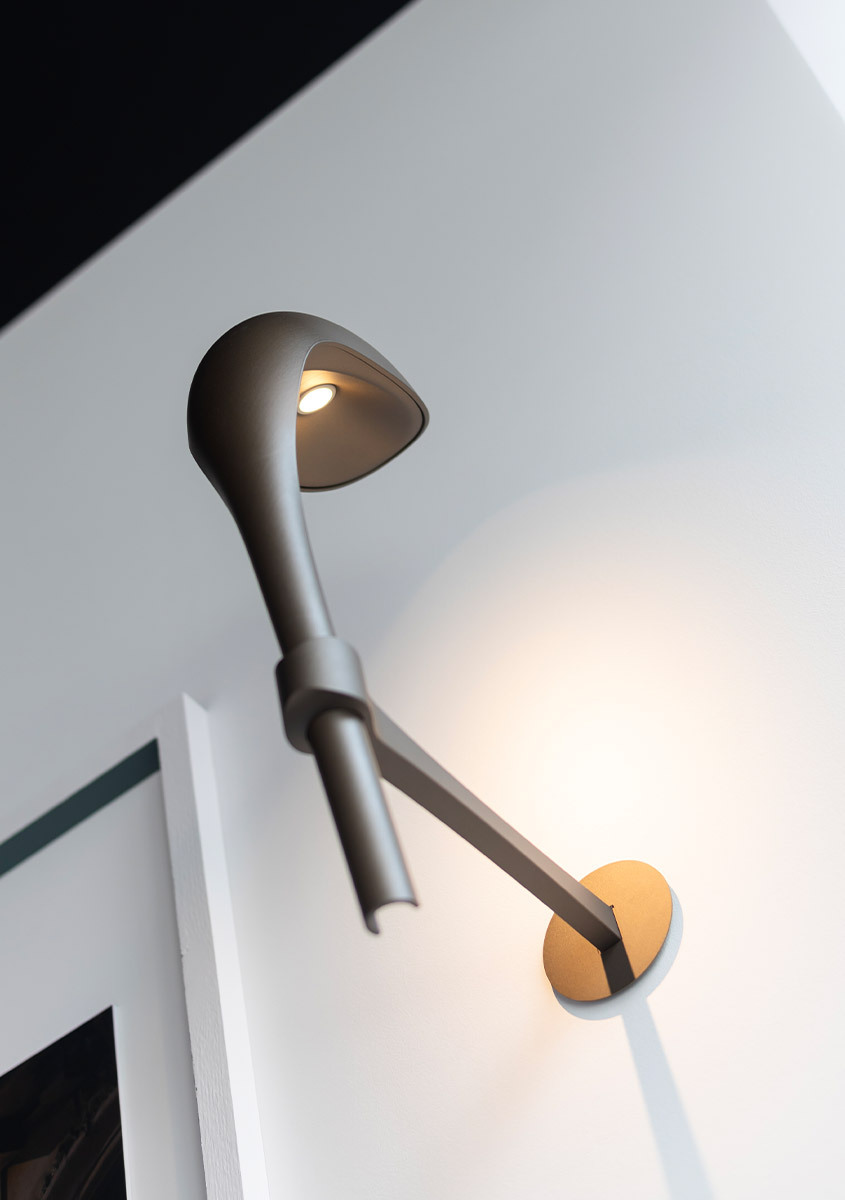 Lampada da parete Artemide con braccio allungato scuro, paralume conico rivolto verso l'alto con luce LED, montata su base circolare dorata a parete