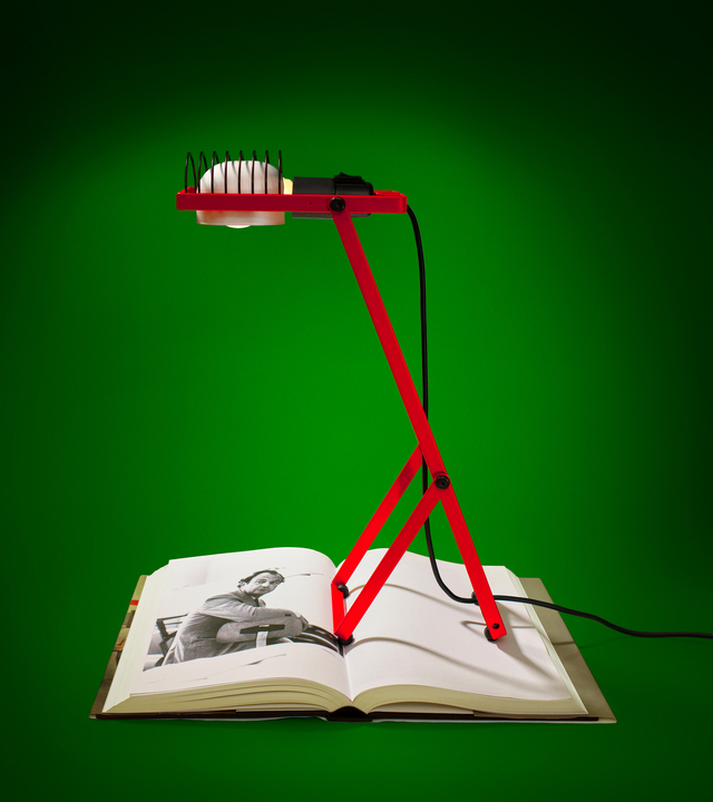 Lampada Sintesi rossa posizionata sopra un catalogo aperto che mostra una foto in bianco e nero di Ernesto Gismondi, su sfondo verde, fotografata da Pierpaolo Ferrari.