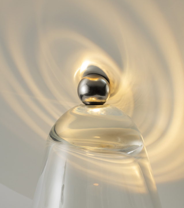 Primo piano della sommità di una lampada in vetro trasparente con finale sferico cromato, che proietta cerchi concentrici di luce sul soffitto