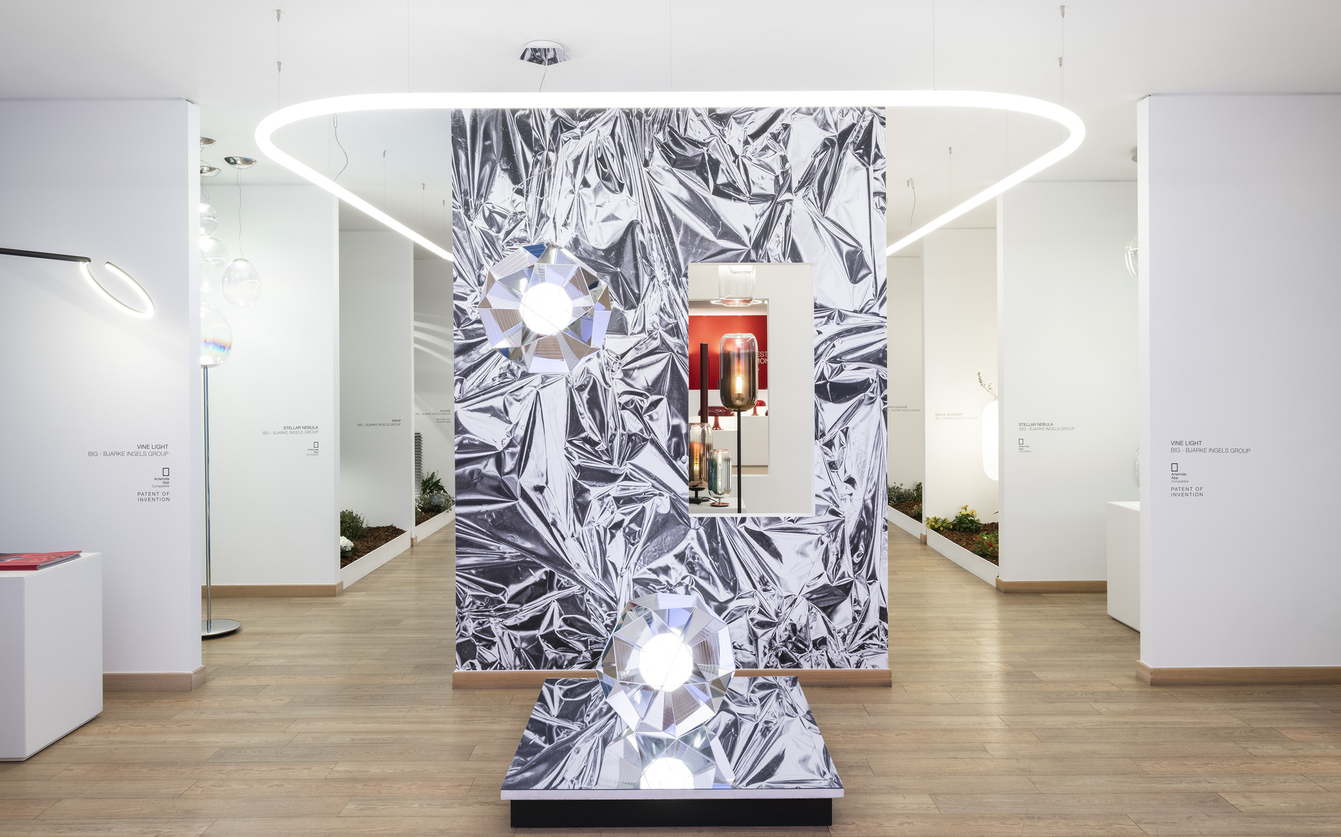 Showroom Artemide con sospensione ad anello LED ovale e due lampade da tavolo sfaccettate su sfondo metallico argentato