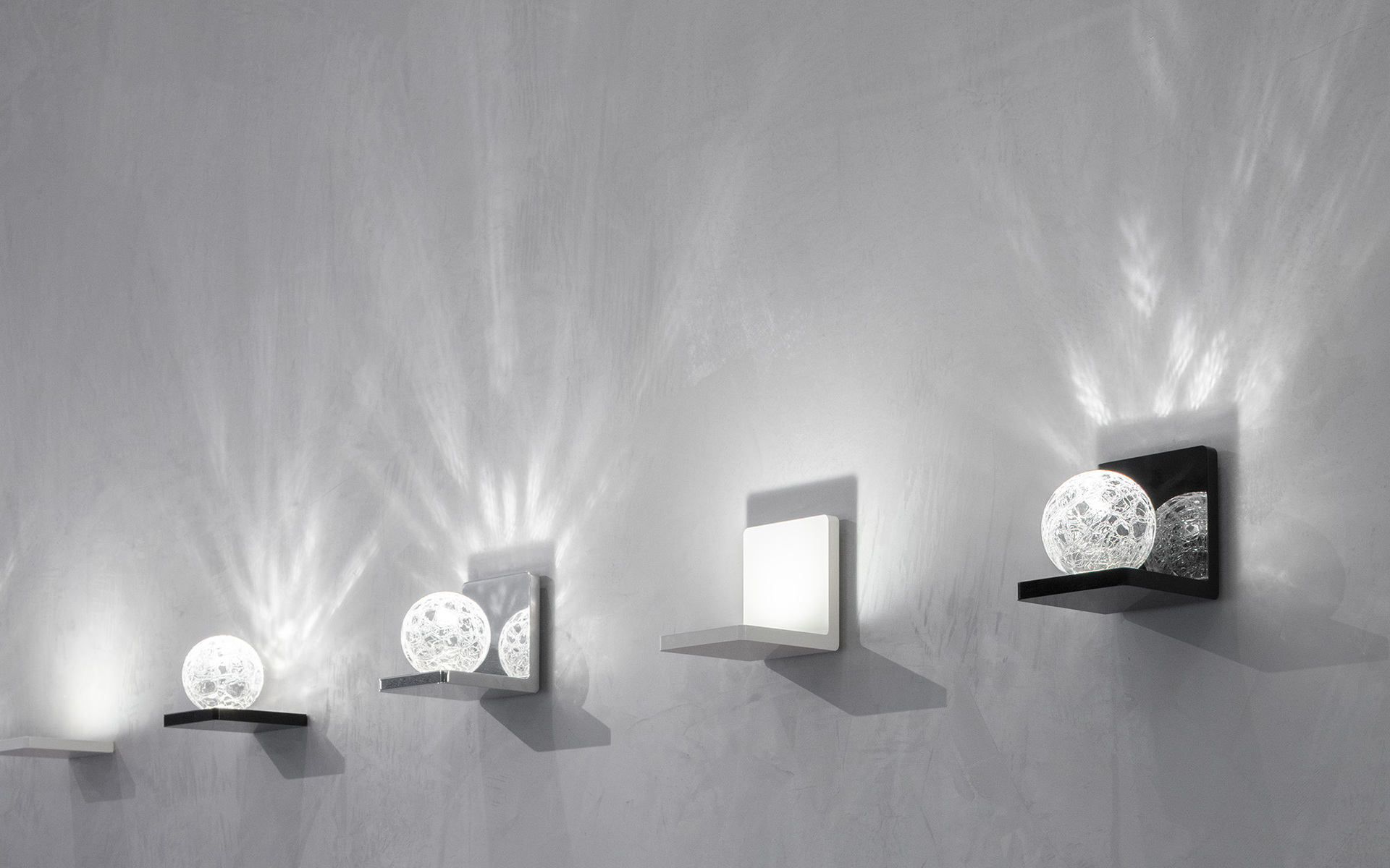 Quattro applique Artemide montate su parete grigia, con diffusori sferici a rete e quadrati che proiettano suggestivi fasci di luce radiali.